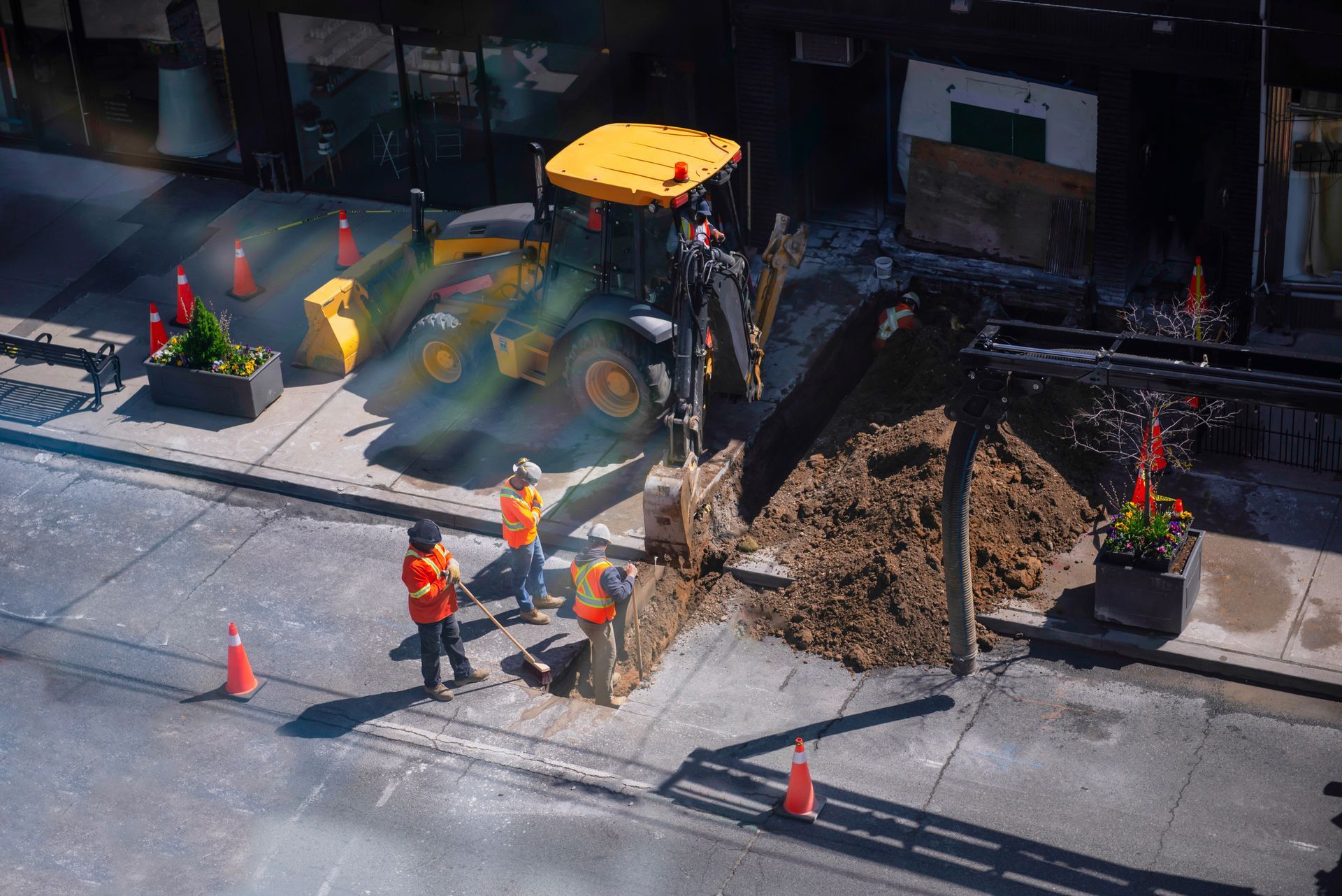 Three construction workers in orange vests use tools near a yellow backhoe at a street excavation site.