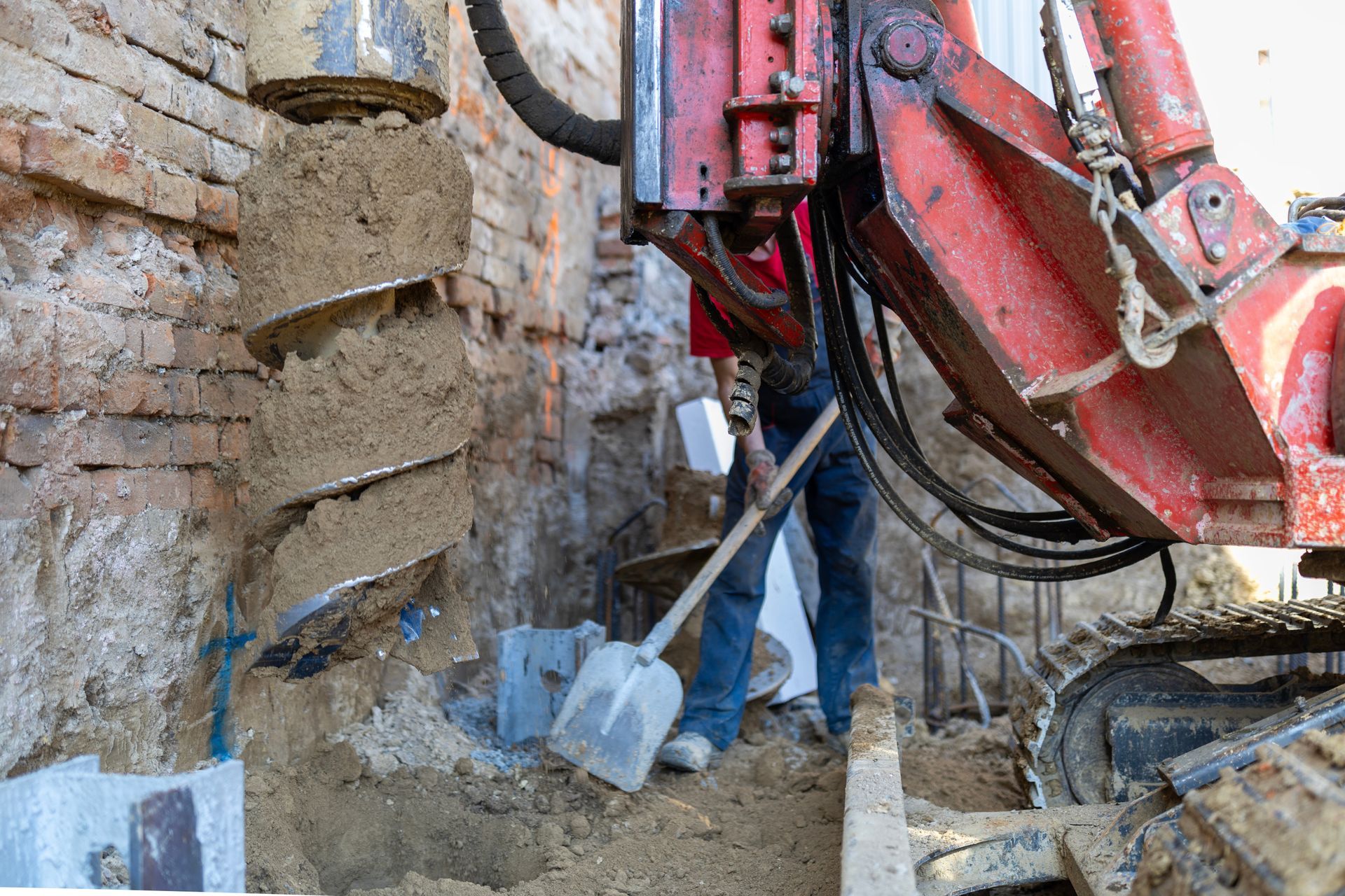 A drilling machine boring a hole into the ground next to a weathered brick wall as a worker holds a shovel nearby.