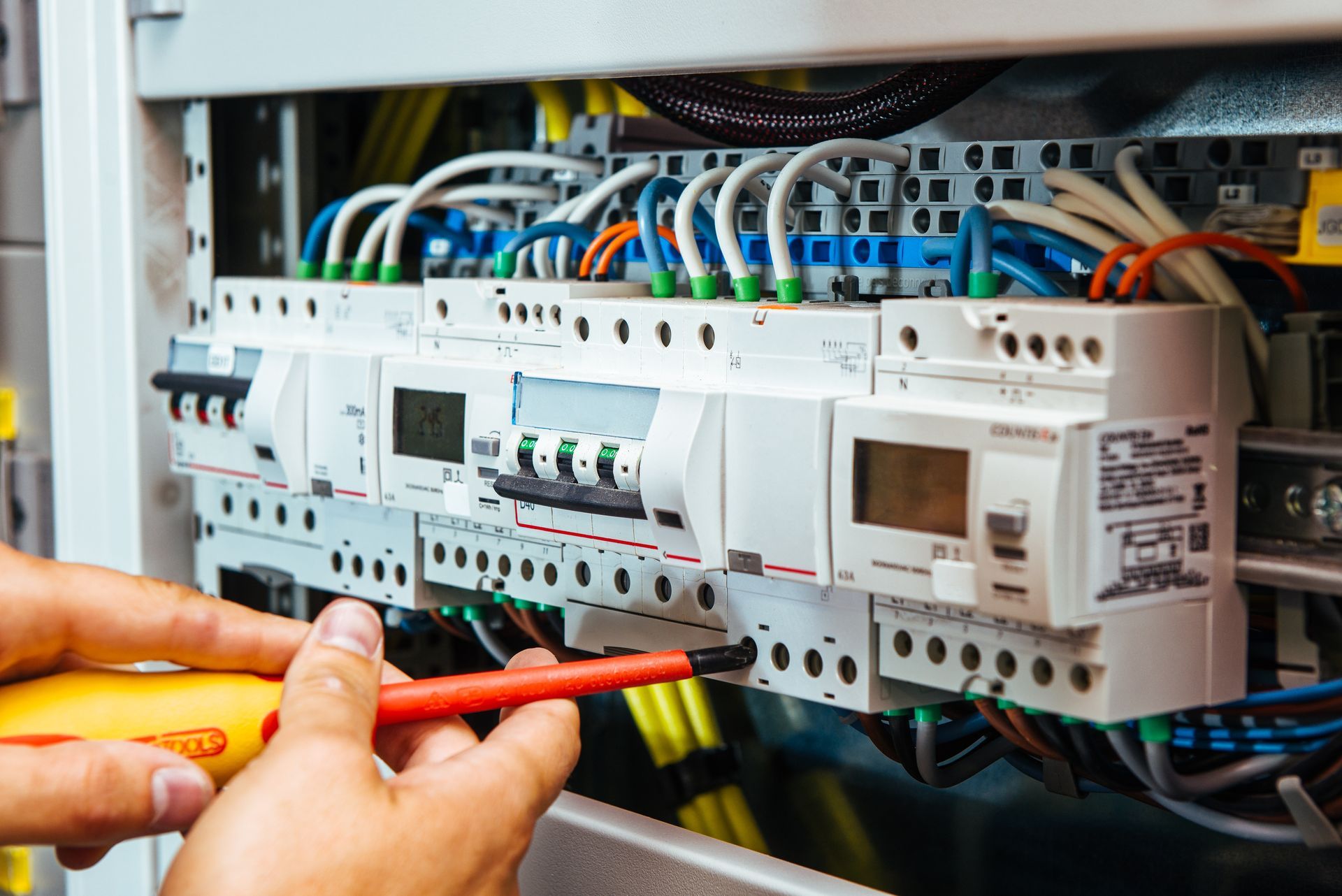Hands using a screwdriver to tighten wires on an electrical circuit breaker panel inside a distribution cabinet.
