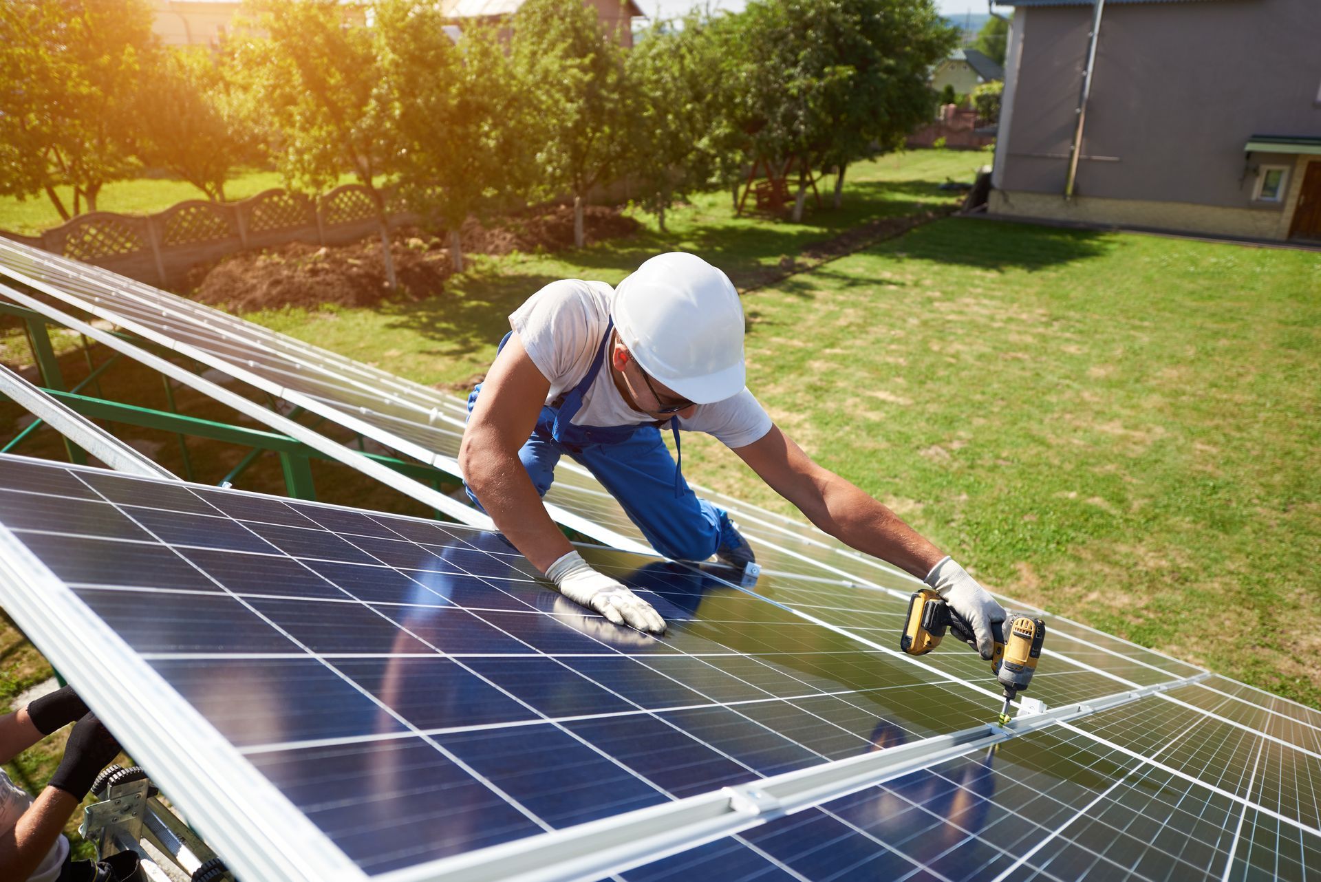 A worker in a white hard hat and blue overalls installs solar panels on a roof in a sunny outdoor setting.