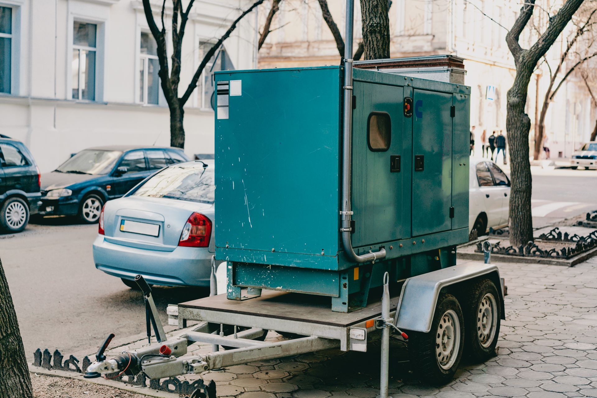 A teal industrial generator on a towable trailer parked on a city street near several cars.