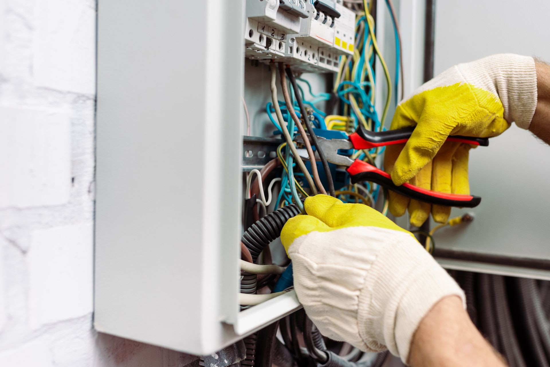 A person wearing yellow and white work gloves uses red-handled pliers to work on electrical wiring in a breaker panel.