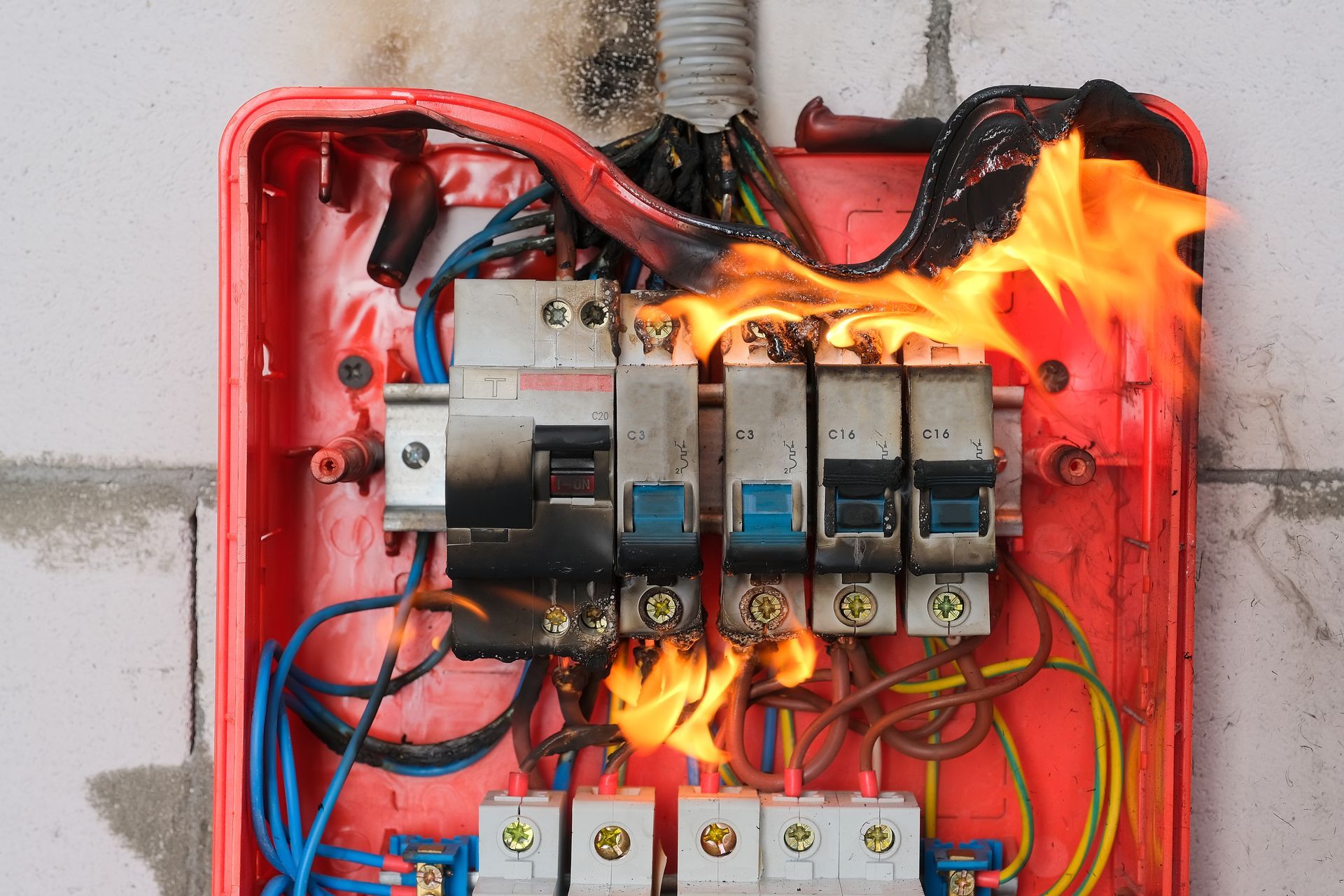 A red electrical breaker box mounted on a wall is actively on fire, with flames engulfing the wiring and circuit breakers.