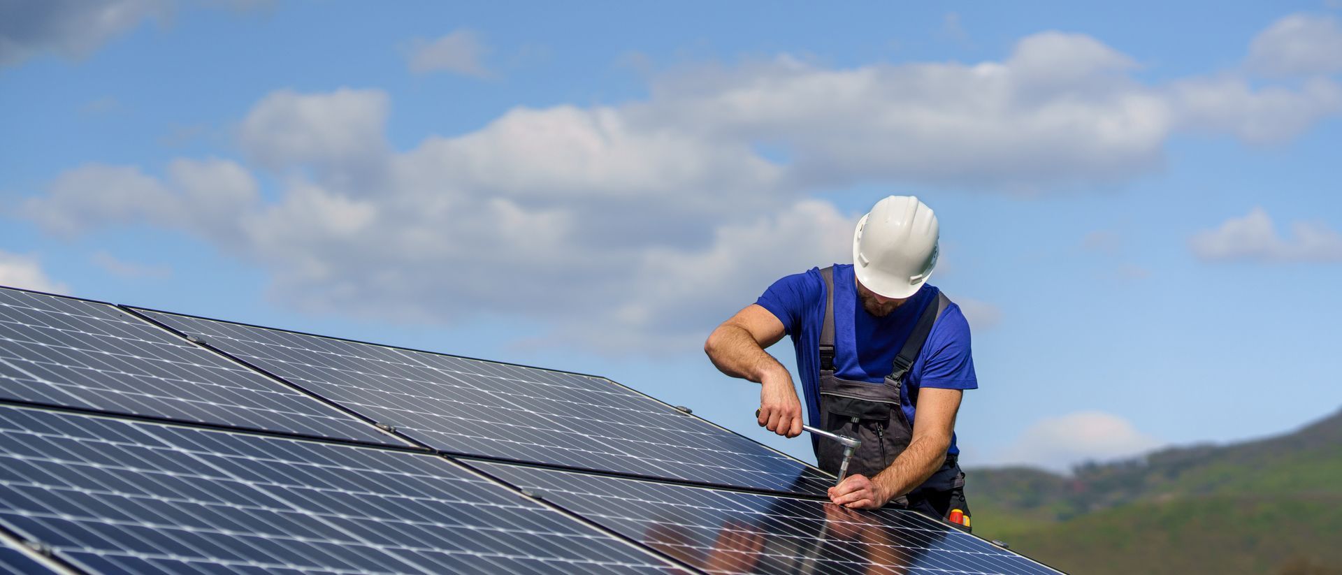 A construction worker in a white hard hat and blue shirt installs solar panels under a partly cloudy sky.