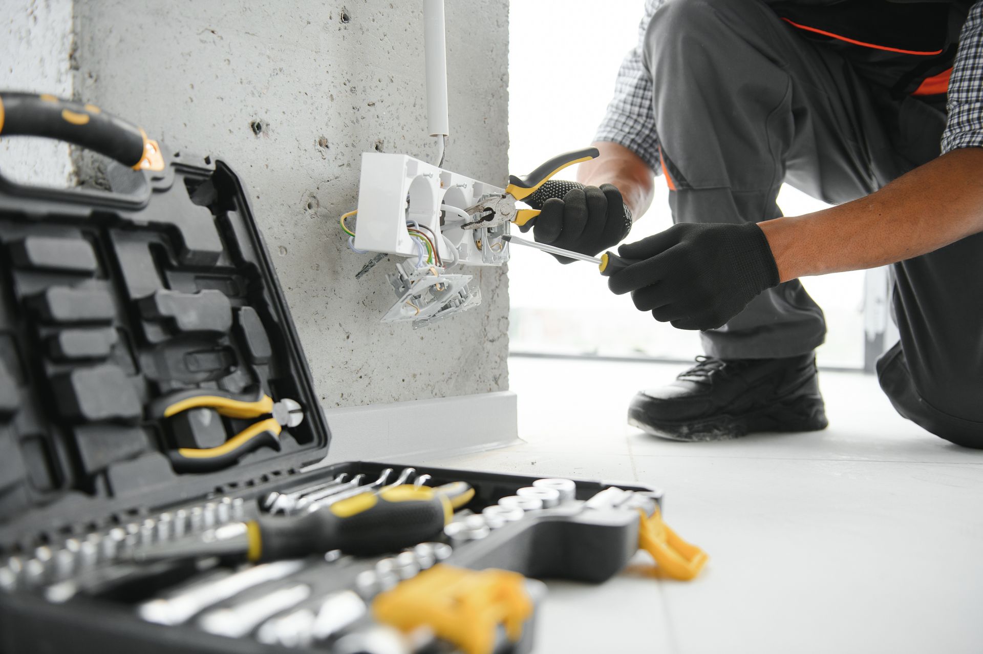 An electrician in black gloves works on a wall-mounted electrical outlet next to an open tool kit on a white floor.