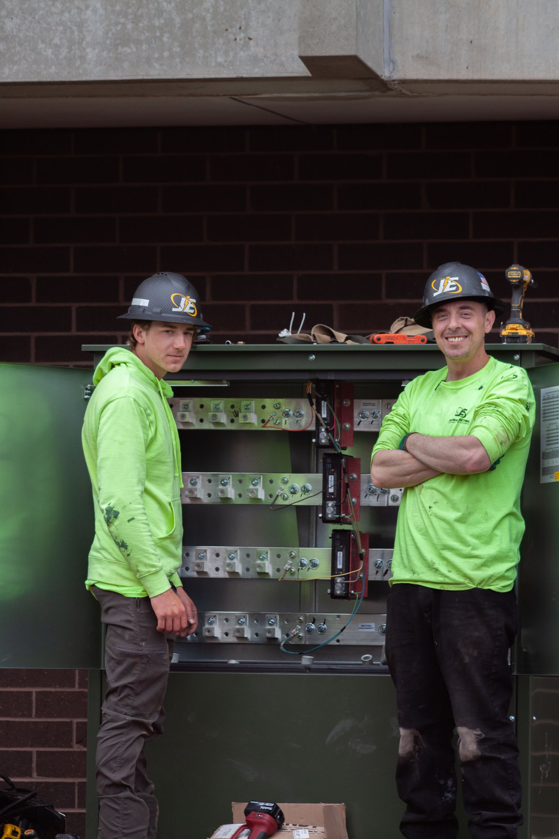 Two utility workers in neon shirts and hard hats standing by an open electrical cabinet against a brick building.