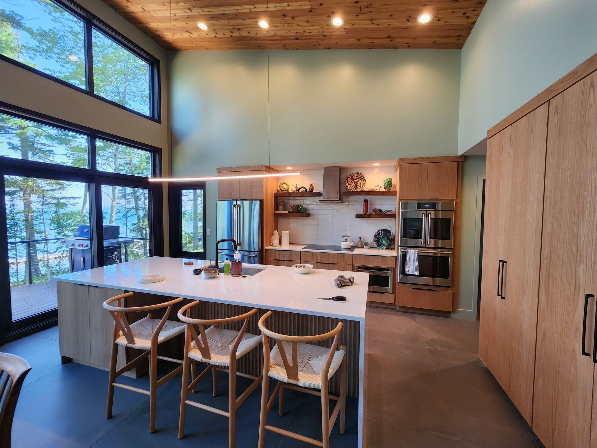 A modern kitchen with light wood cabinets, a large white island with three wooden stools, and tall windows facing trees.