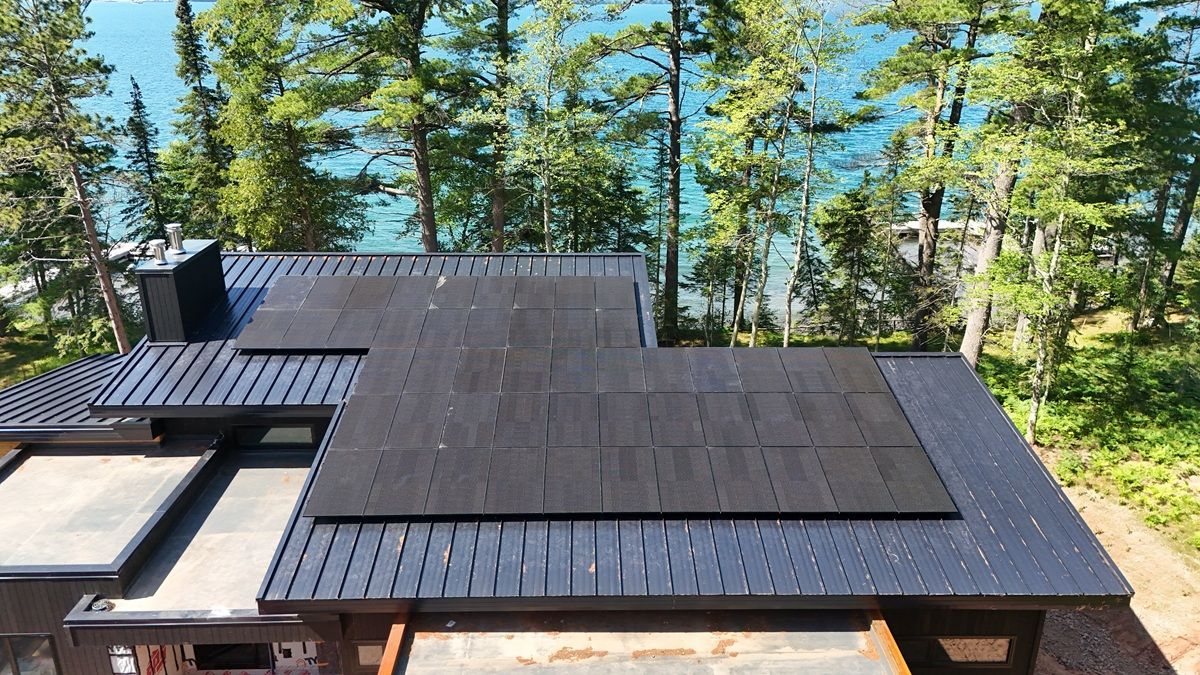 Aerial view of a black metal roof with a large, integrated solar panel array surrounded by trees and a lake.