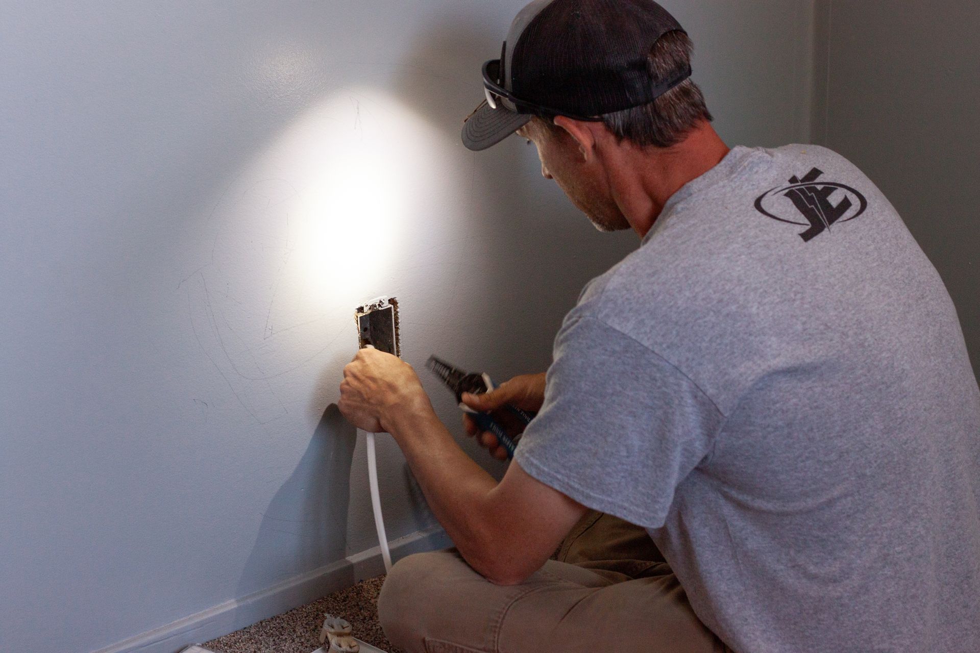 A person wearing a grey t-shirt and headlamp kneels on the floor, using pliers to work on electrical wiring in a wall.