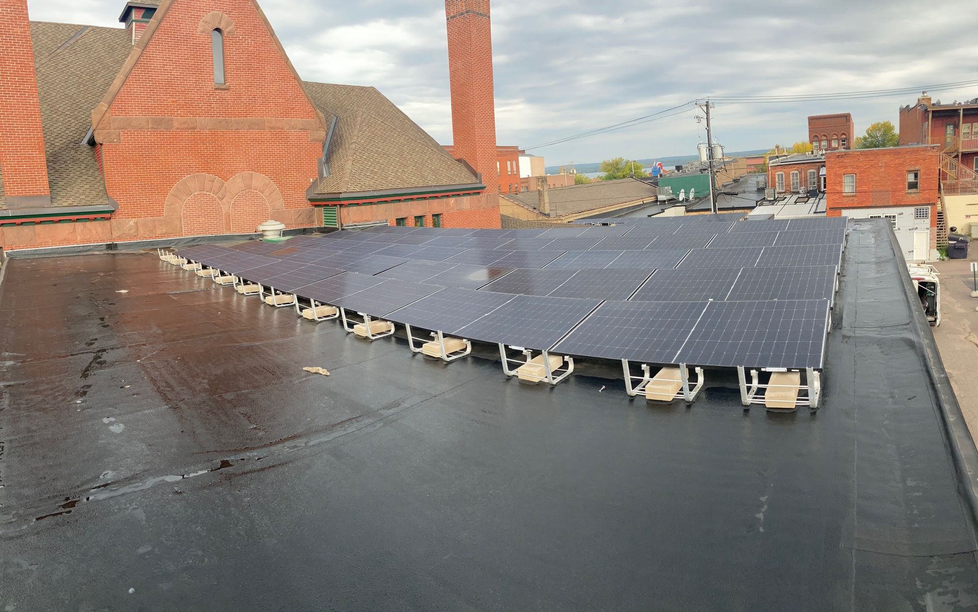 Solar panels mounted on a flat, dark roof in a city with a brick building in the background under a cloudy sky.