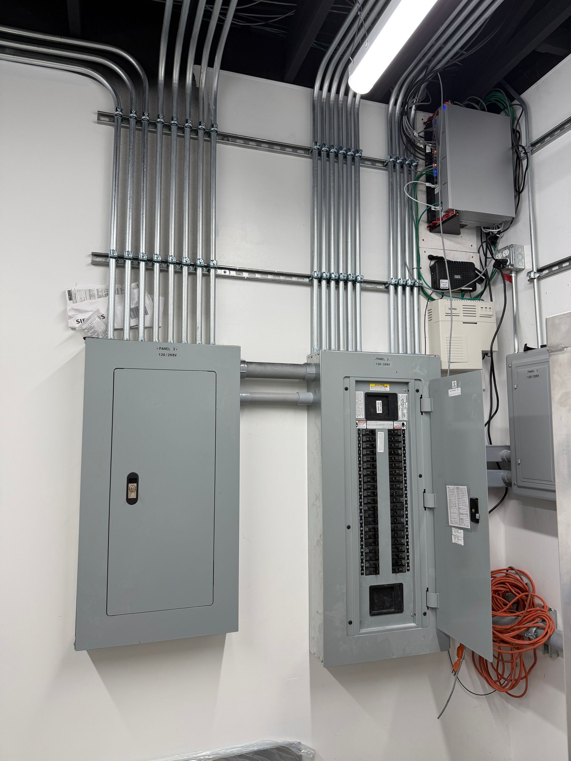 Two grey electrical service panels mounted on a white wall, connected by extensive metal conduit piping overhead.