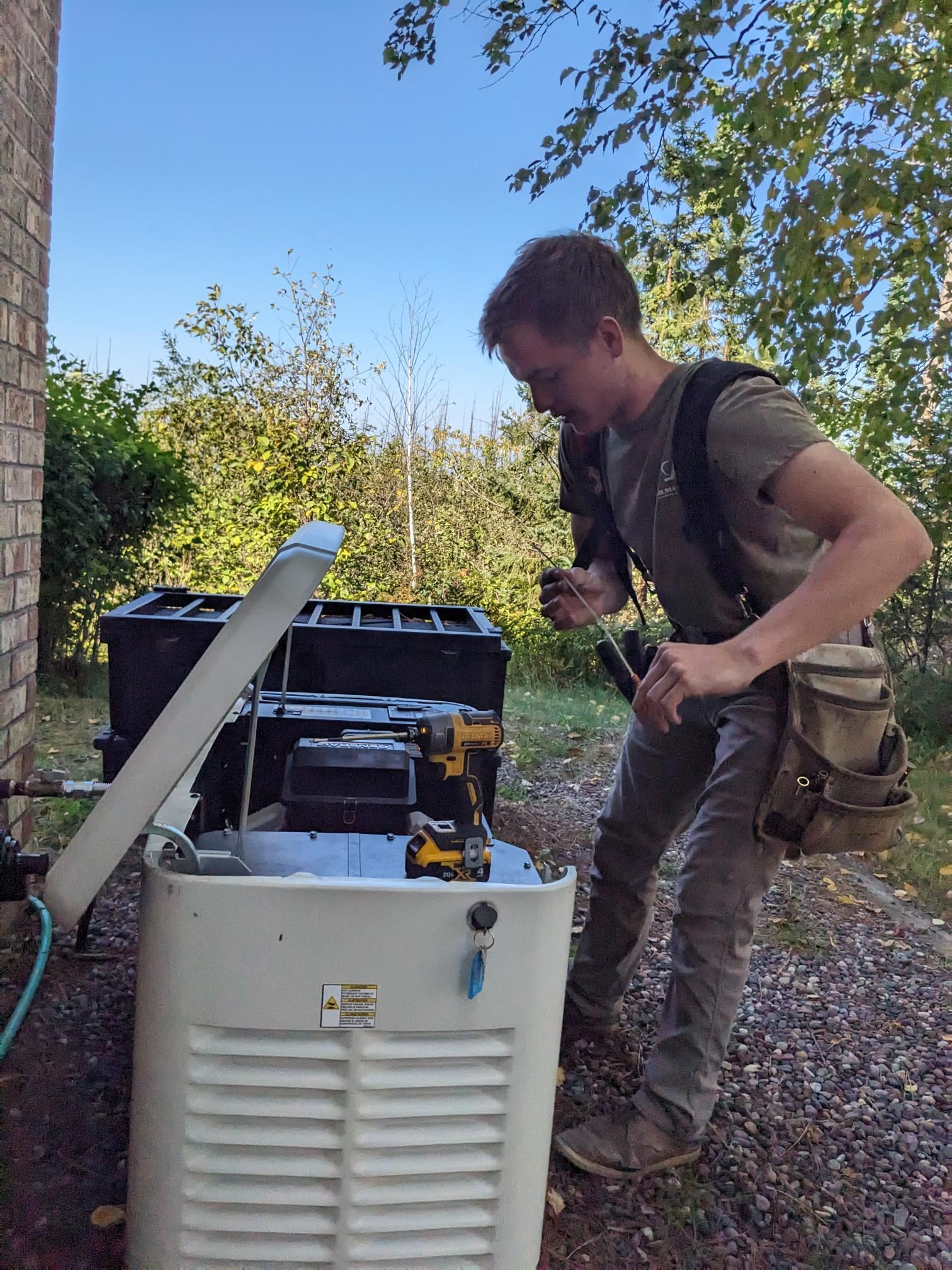 A technician works on an open standby generator outdoors, using a power drill to perform maintenance.