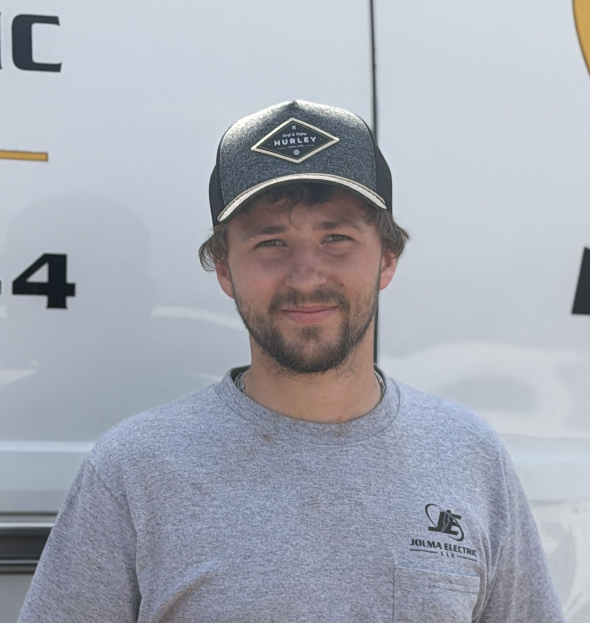 Man in a black cap and gray shirt standing in front of a white vehicle, smiling slightly.