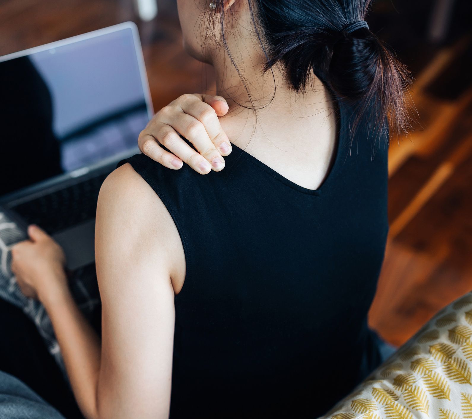 Woman sitting, touching shoulder, using laptop; possible neck/shoulder pain, indoors.