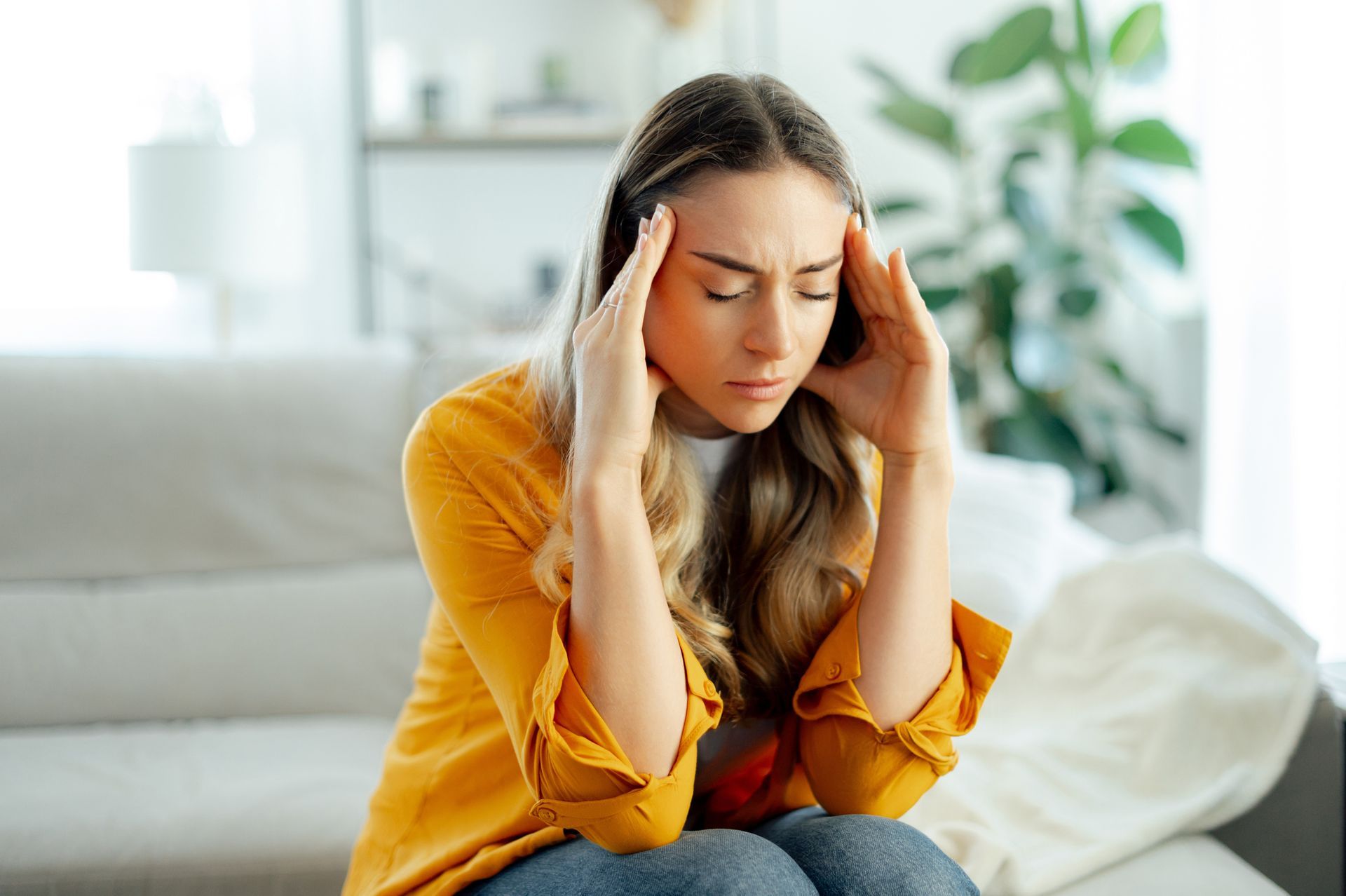Woman with headache, hands on temples, seated on a couch in a living room.