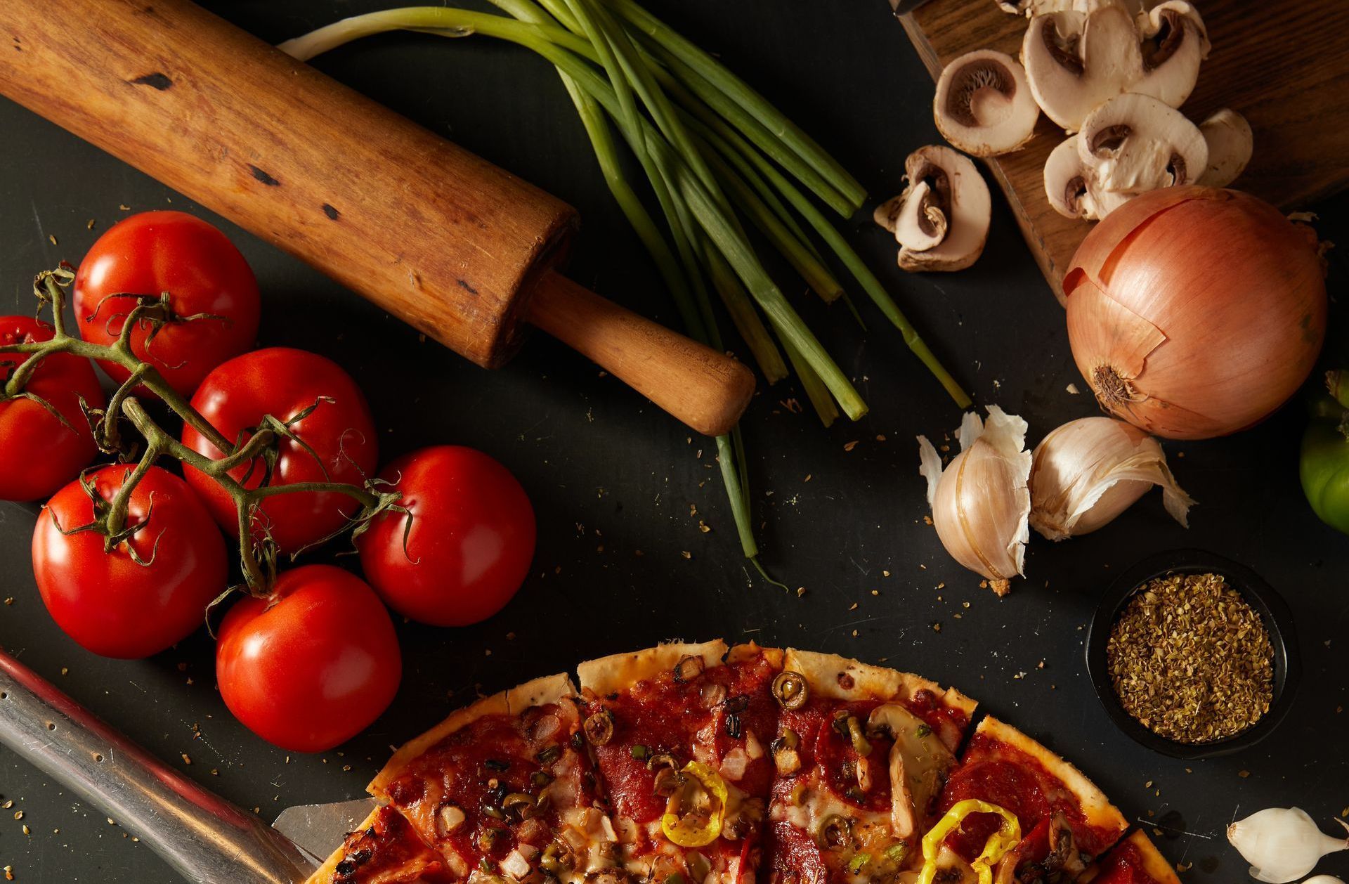 A pizza is surrounded by tomatoes , mushrooms , onions and a rolling pin.