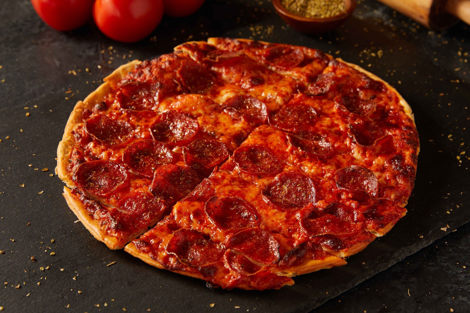 A pepperoni pizza is sitting on a table with tomatoes in the background.