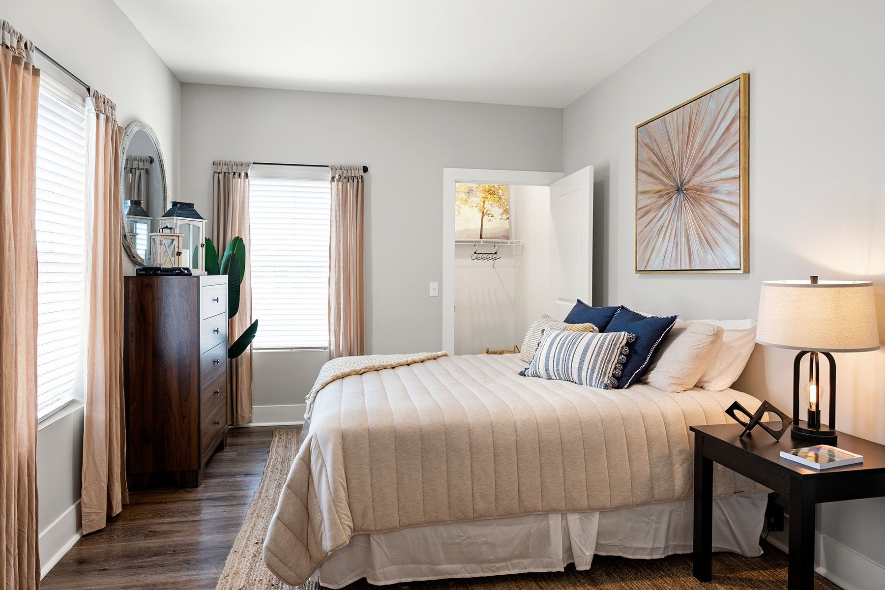 Pointe Grand Columbia Apartment Homes' spacious bedroom features hardwood-style flooring, a bed, wall art, nightstand, dresser, mirror, and plants.
