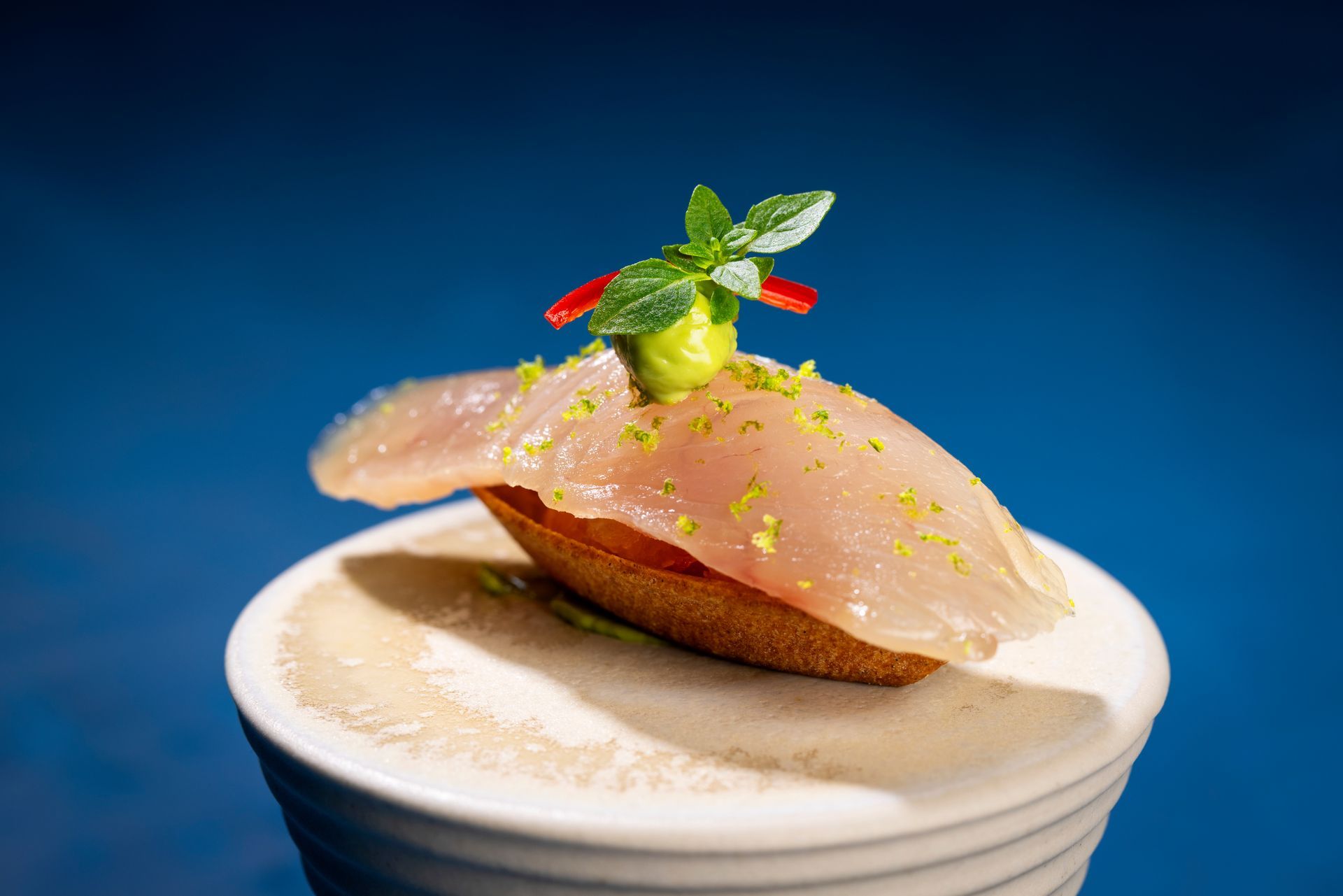 Close-up van een gastronomisch voorgerecht. Een dun plakje vis op geroosterd brood met groene en rode garnering, op een klein wit schaaltje.