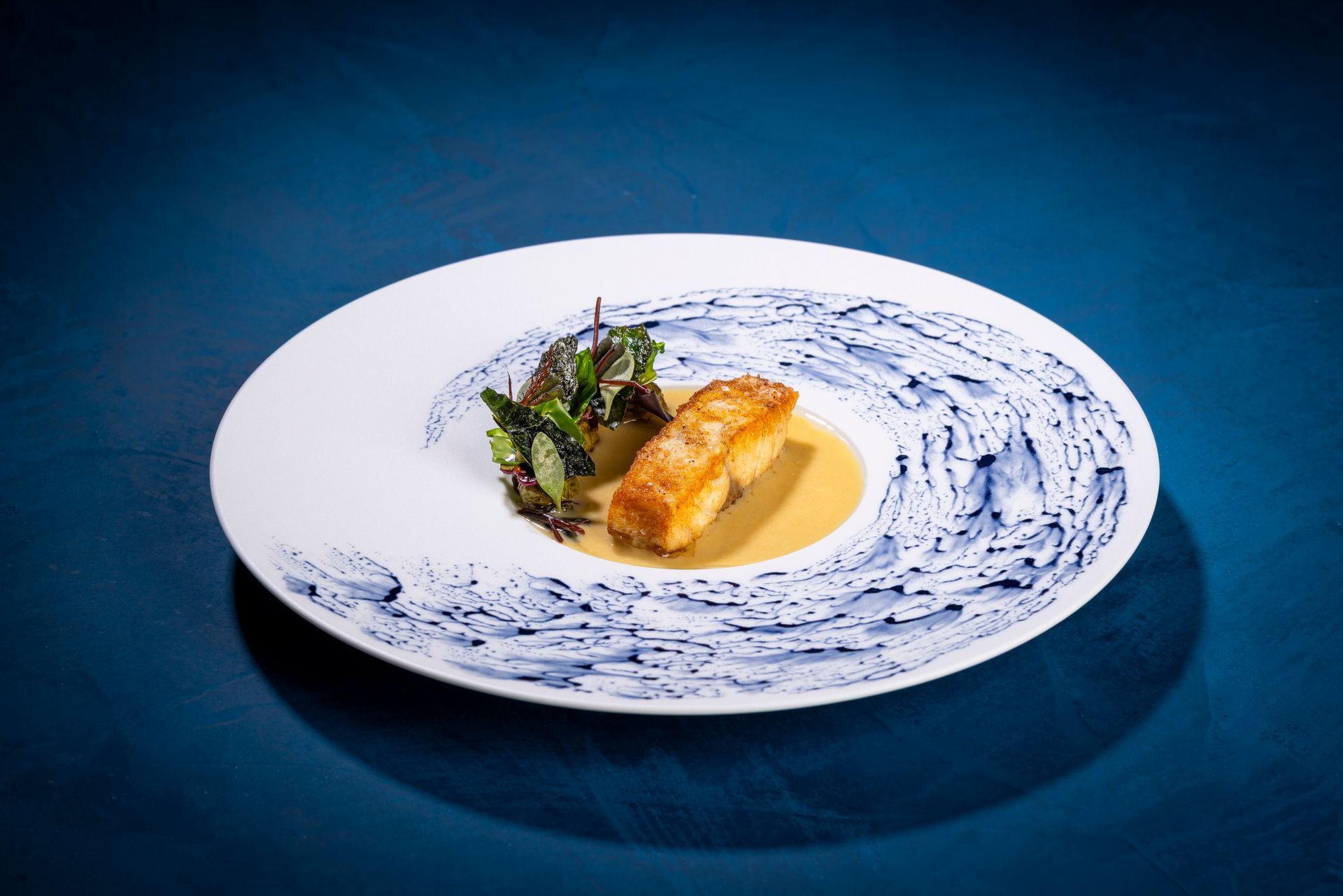 Een bord met eten, een stuk vis, saus en groenten op een blauw bord tegen een blauwe achtergrond.