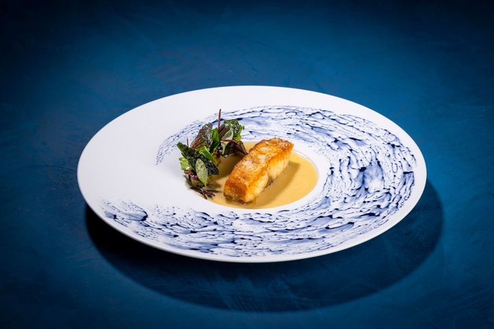 Gourmet fish dish on a swirl-patterned plate with green garnish and sauce on a blue surface.