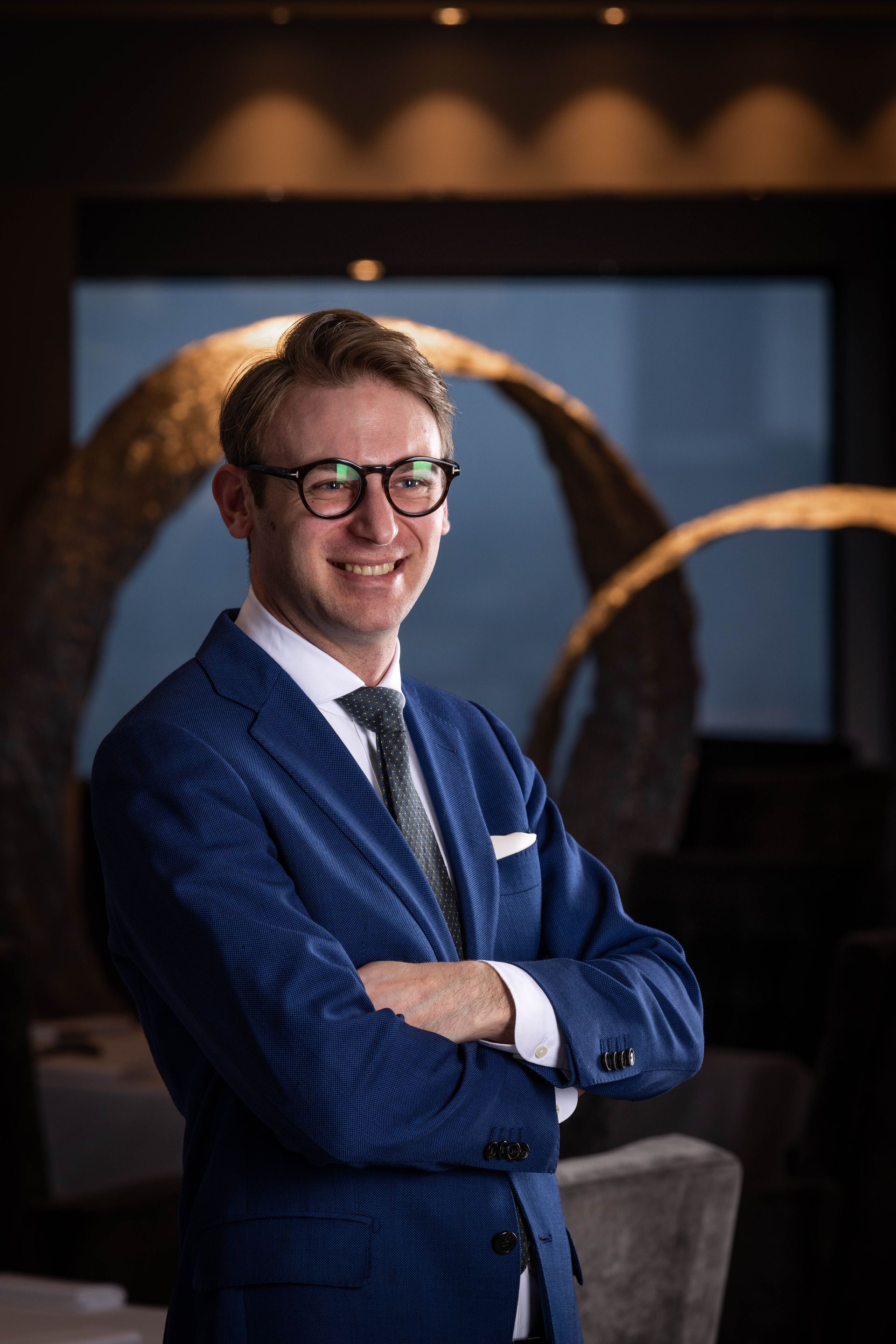 Een man in een blauw pak en stropdas staat met zijn armen over elkaar en lacht.