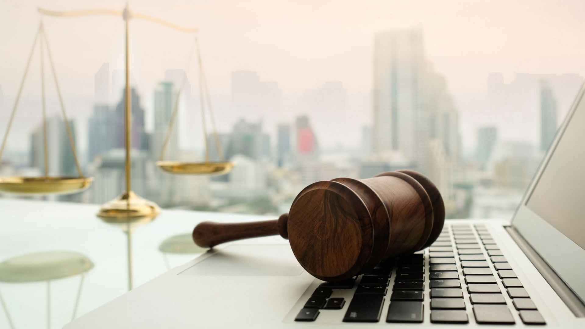 Gavel on laptop keyboard with scales of justice in front of city skyline.