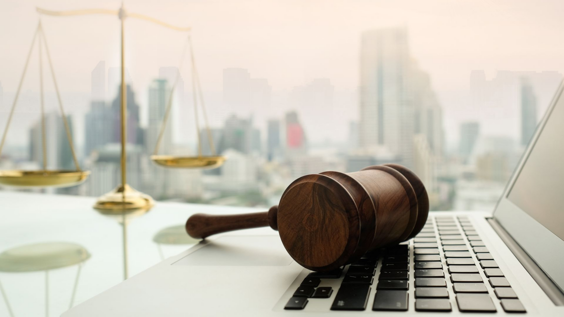 Gavel on laptop keyboard with scales of justice in front of city skyline.