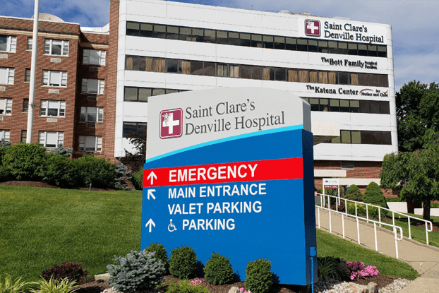 Sign for Saint Clare's Denville Hospital, with directional arrows for Emergency, Main Entrance, and Parking, with hospital building in background.