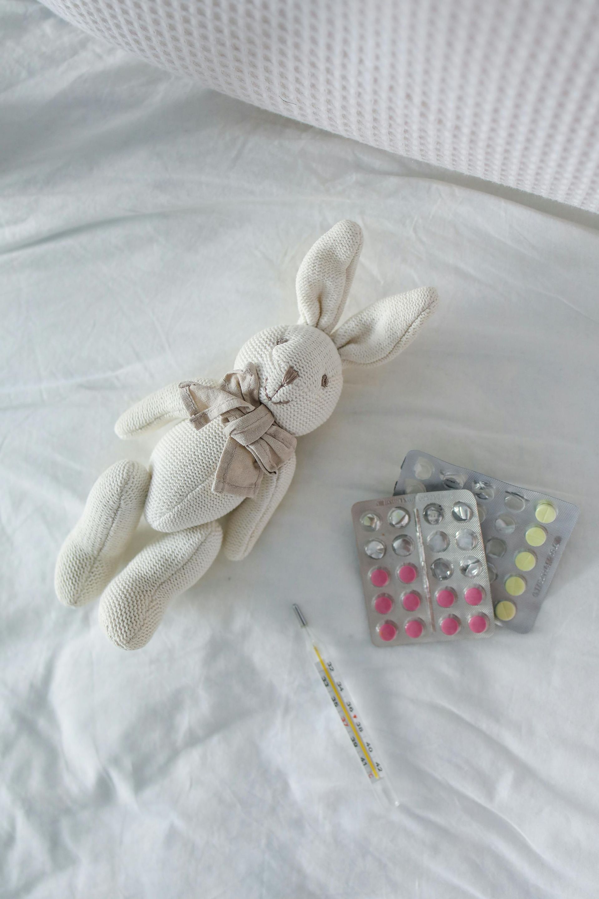 Bunny toy, blister packs of pills, and thermometer on white bedsheets.