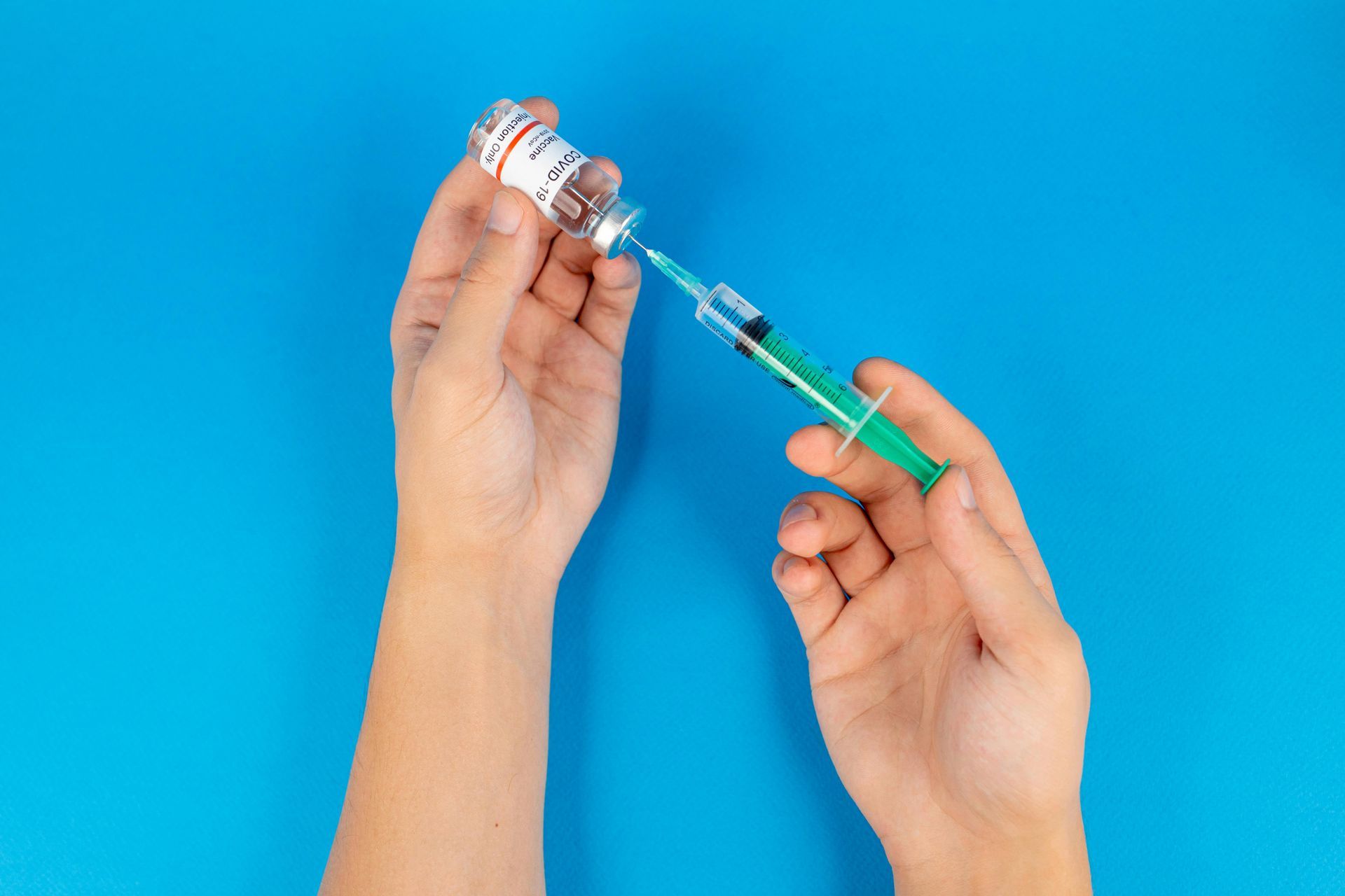 Hands filling a syringe from a vaccine vial against a blue background.