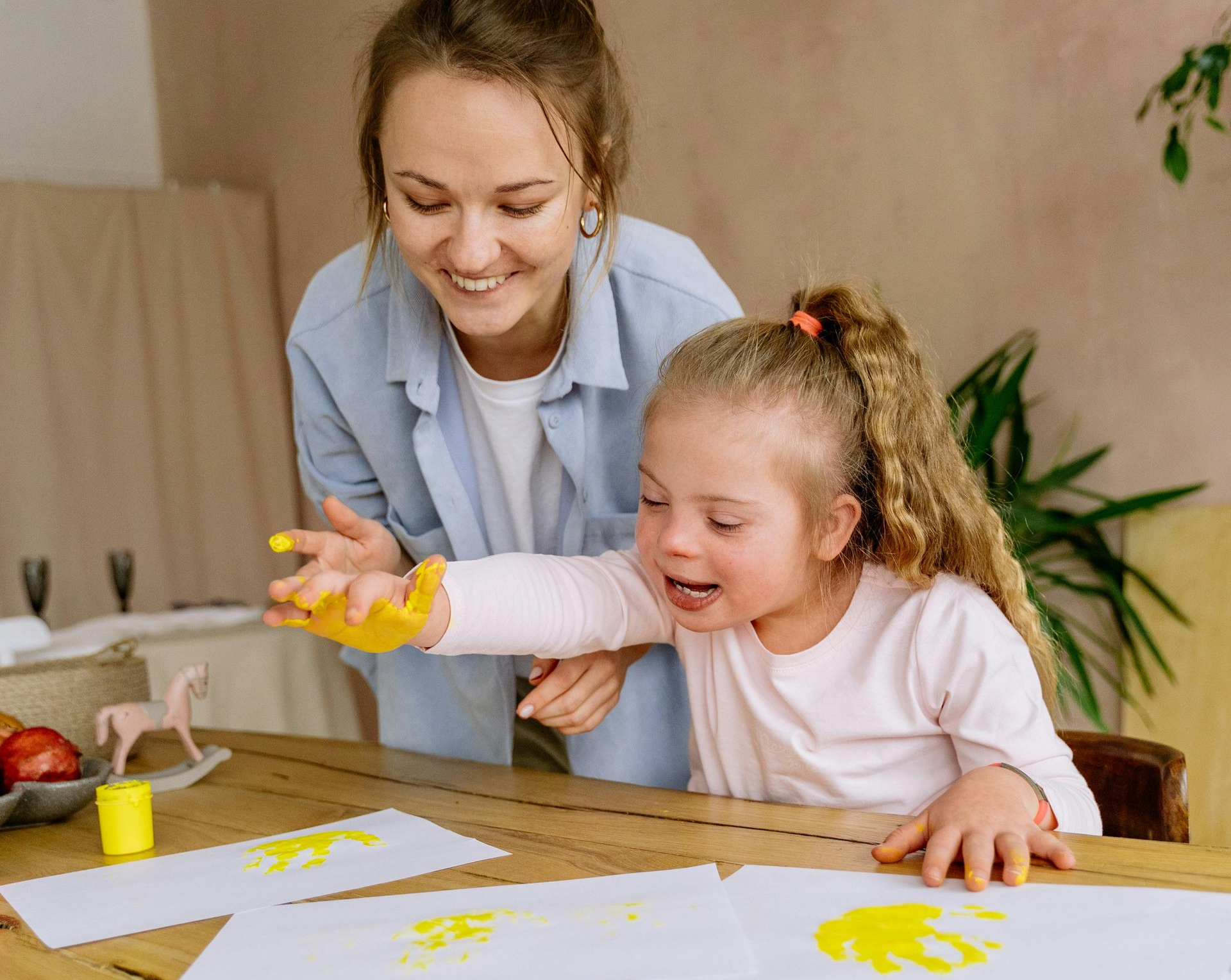 Woman helps child with Down syndrome paint with yellow paint at a table.