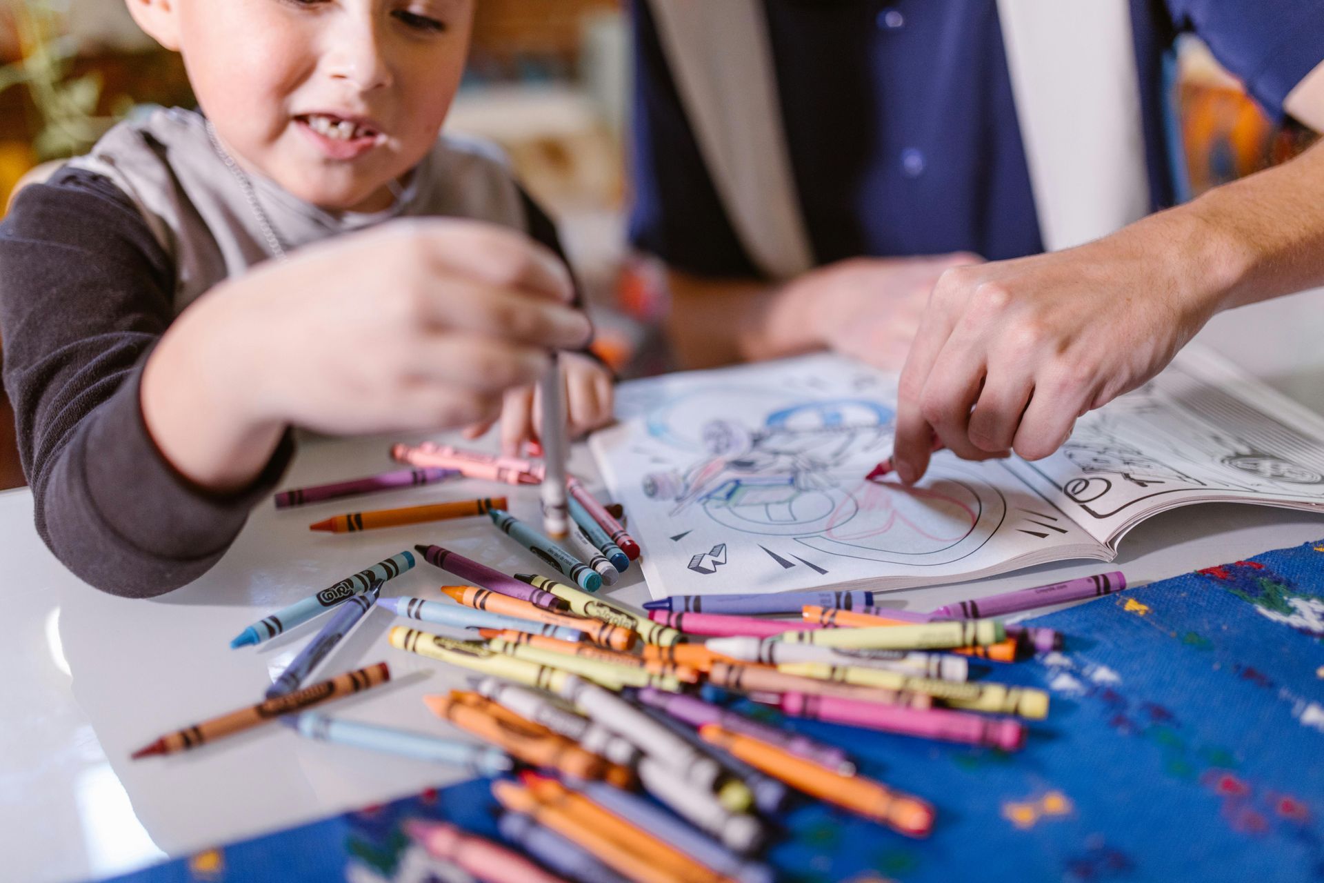 Child coloring in a book with crayons, adult's hand pointing to a page. Crayons scattered on a table.