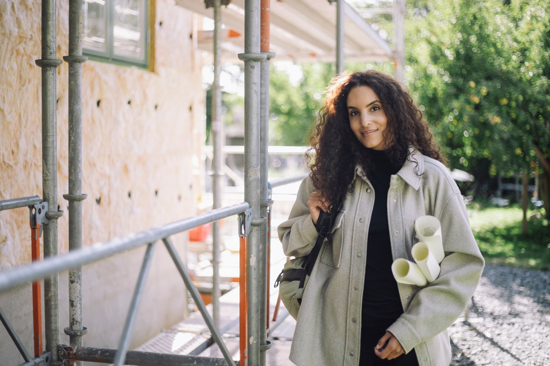 A woman is standing in front of a building under construction holding blueprints.