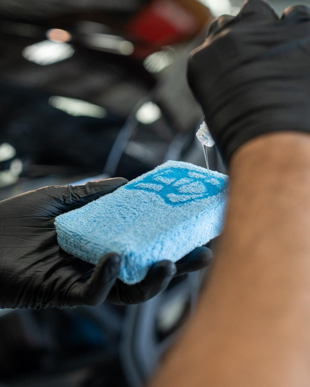 Hands in black gloves applying liquid to a blue applicator pad near a car.