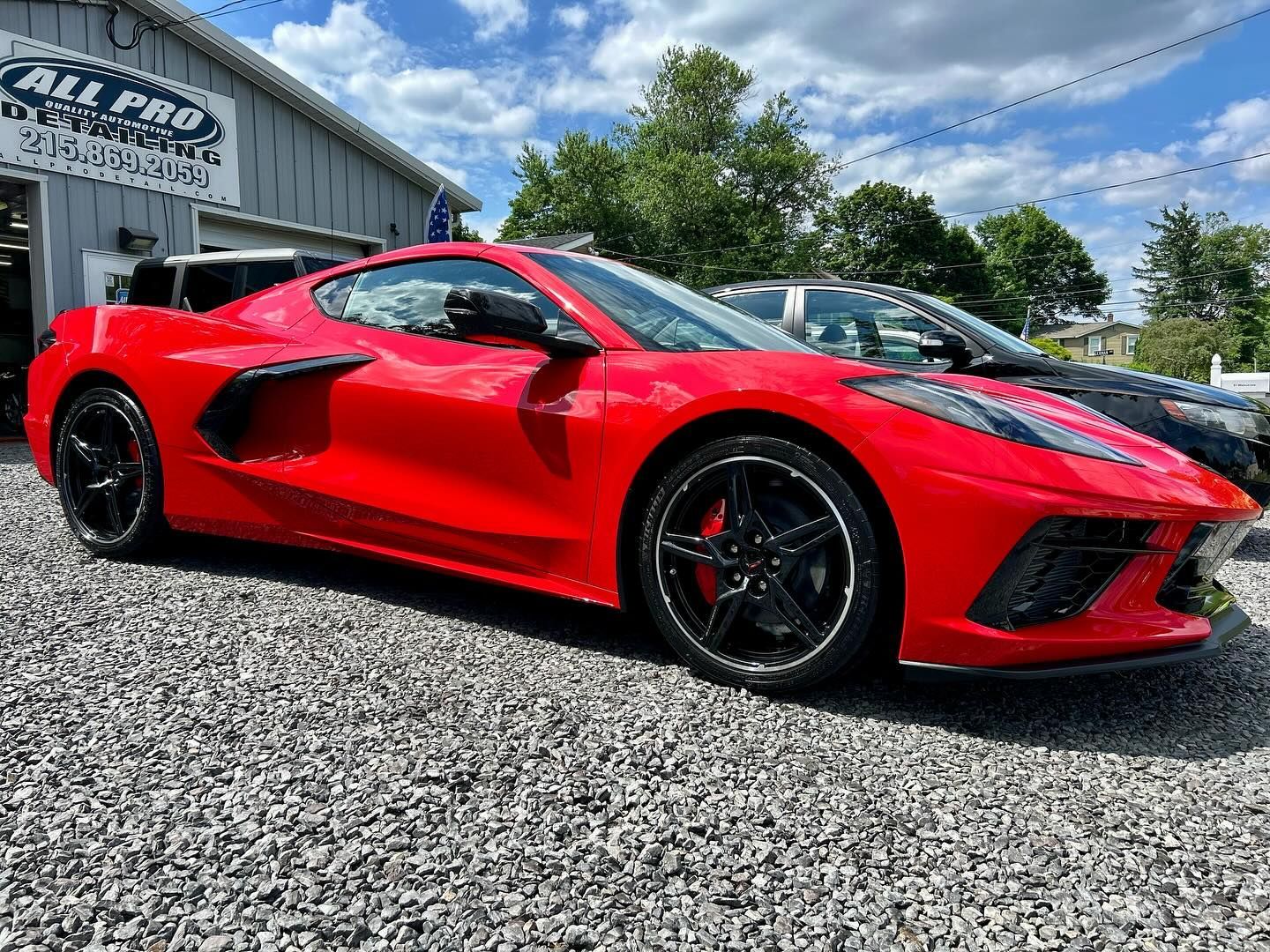 A bright red Corvette sports car parked on a gravel lot in front of a building under a blue sky with scattered clouds.