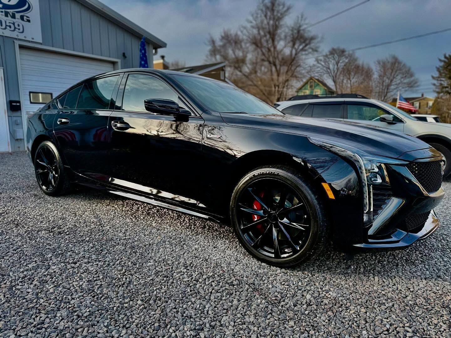 A glossy black Cadillac CT4-V sedan parked on a gravel lot in front of a building.