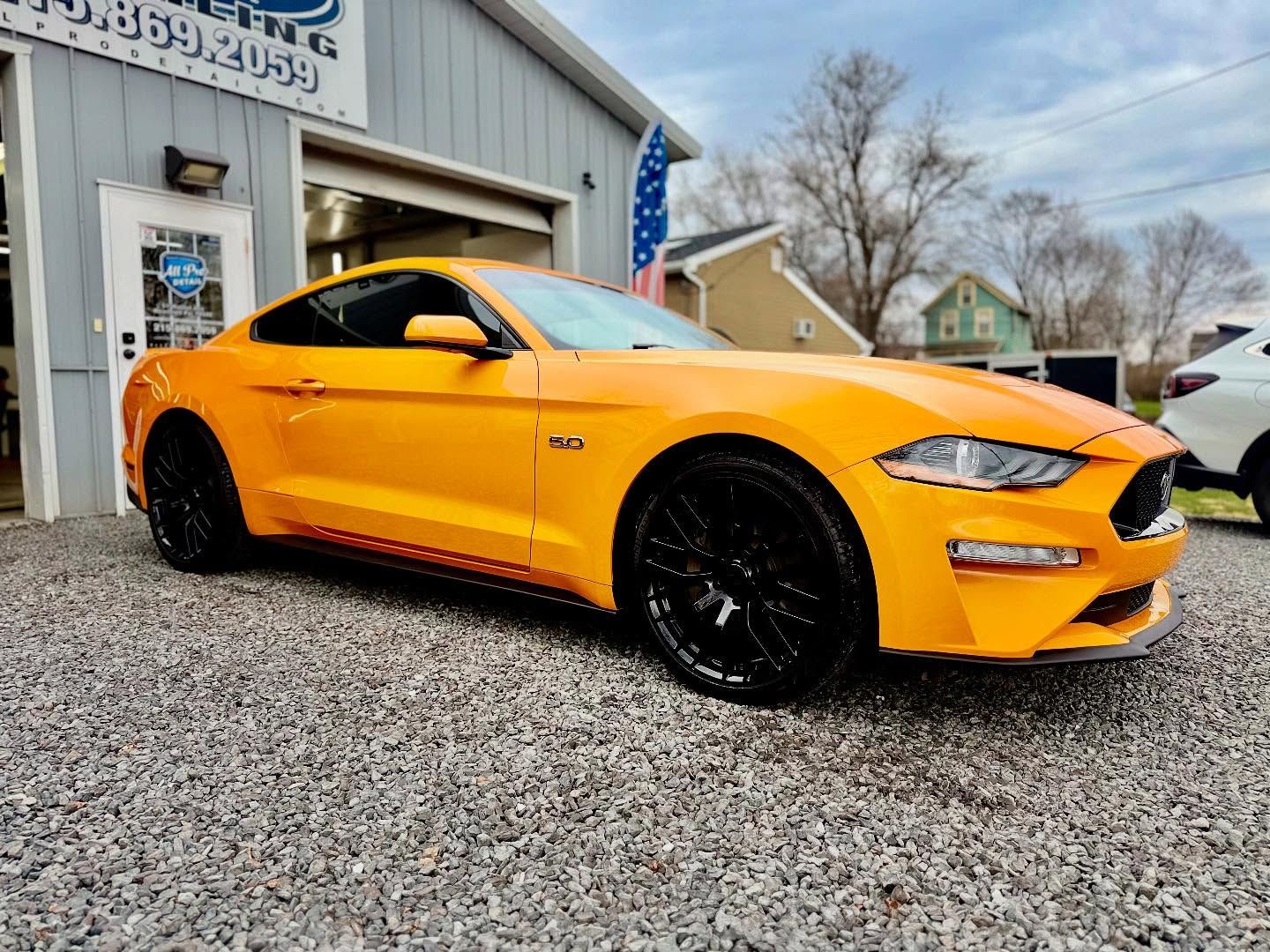 A bright orange Ford Mustang parked on a gravel lot in front of a building with an American flag.