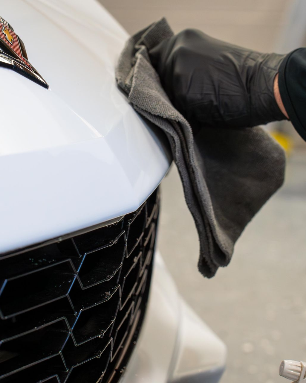 Gloved hand wiping a white car's hood with a gray microfiber cloth; close-up view.