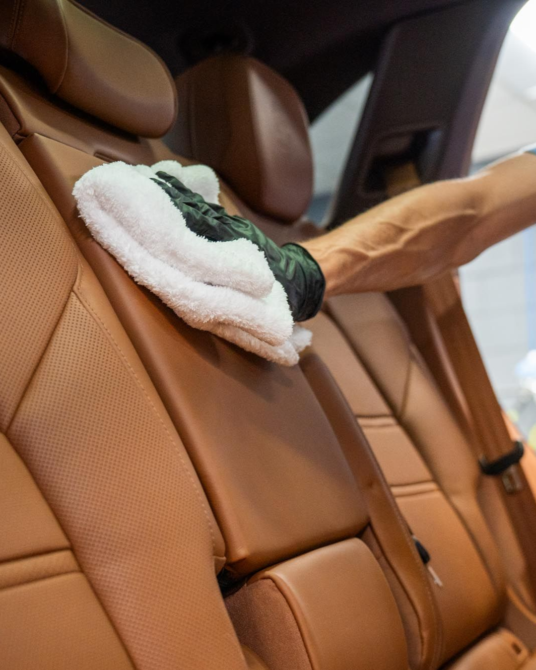 Hand in black glove wiping tan leather car seat with white cloth.