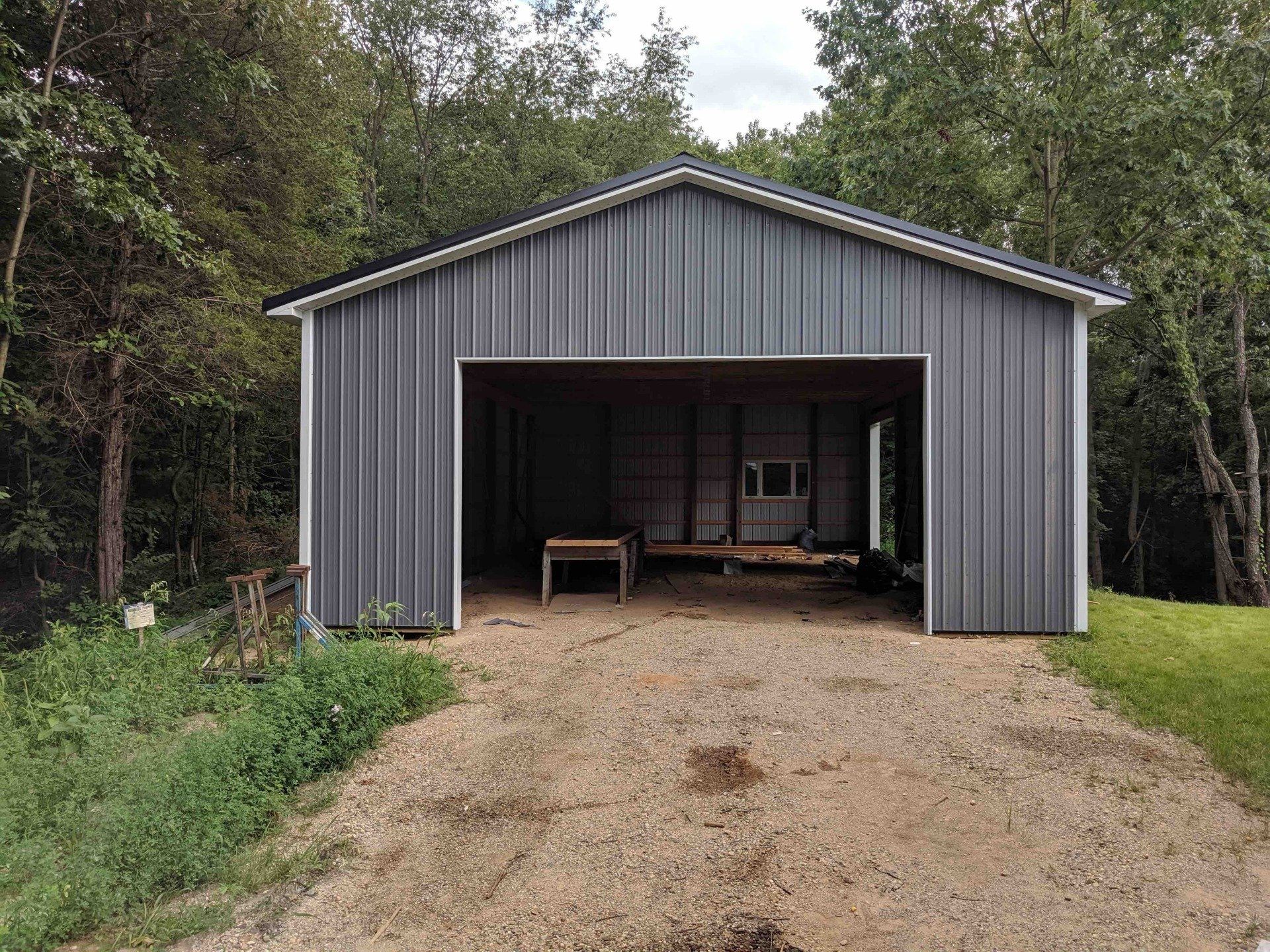 a gray metal garage is sitting on top of a dirt road in the middle of the woods.