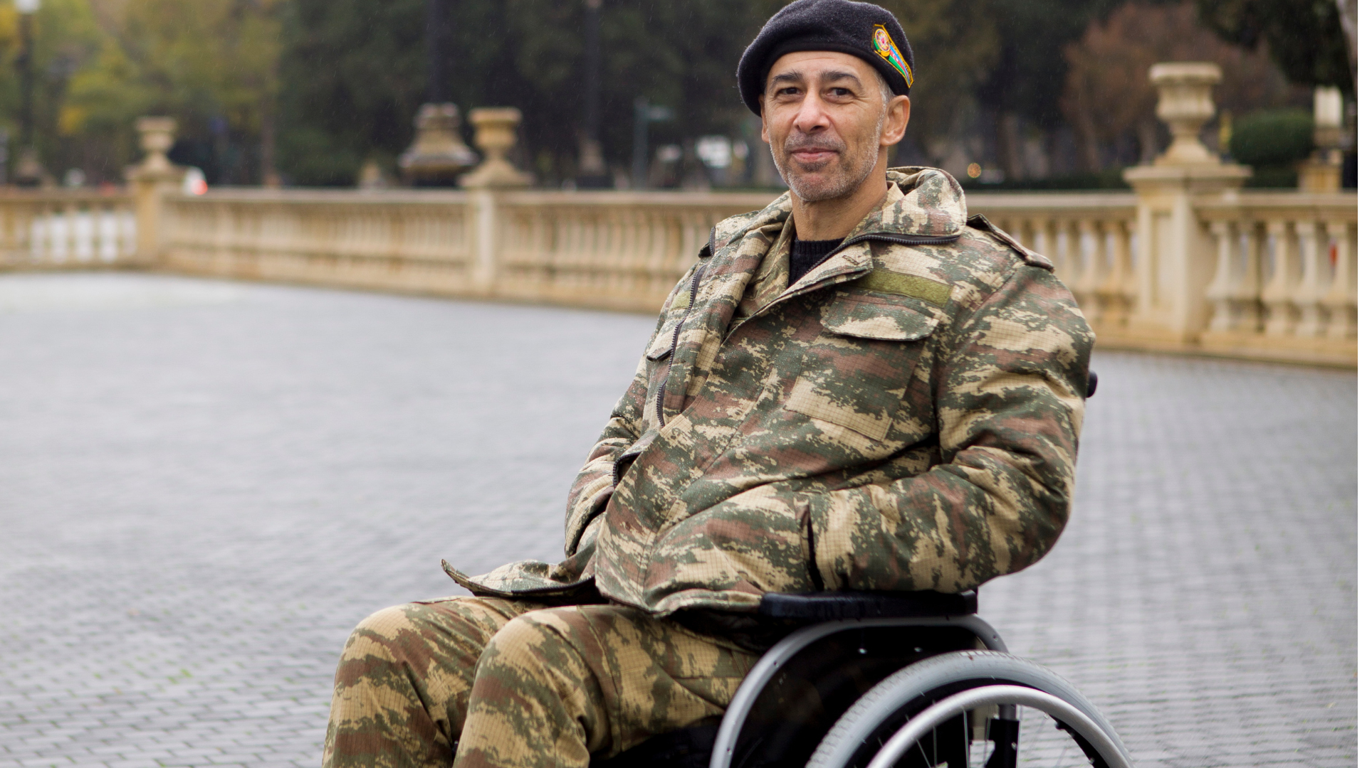 Man in camouflage uniform and beret in a wheelchair smiles. Outdoors setting with stone architecture.
