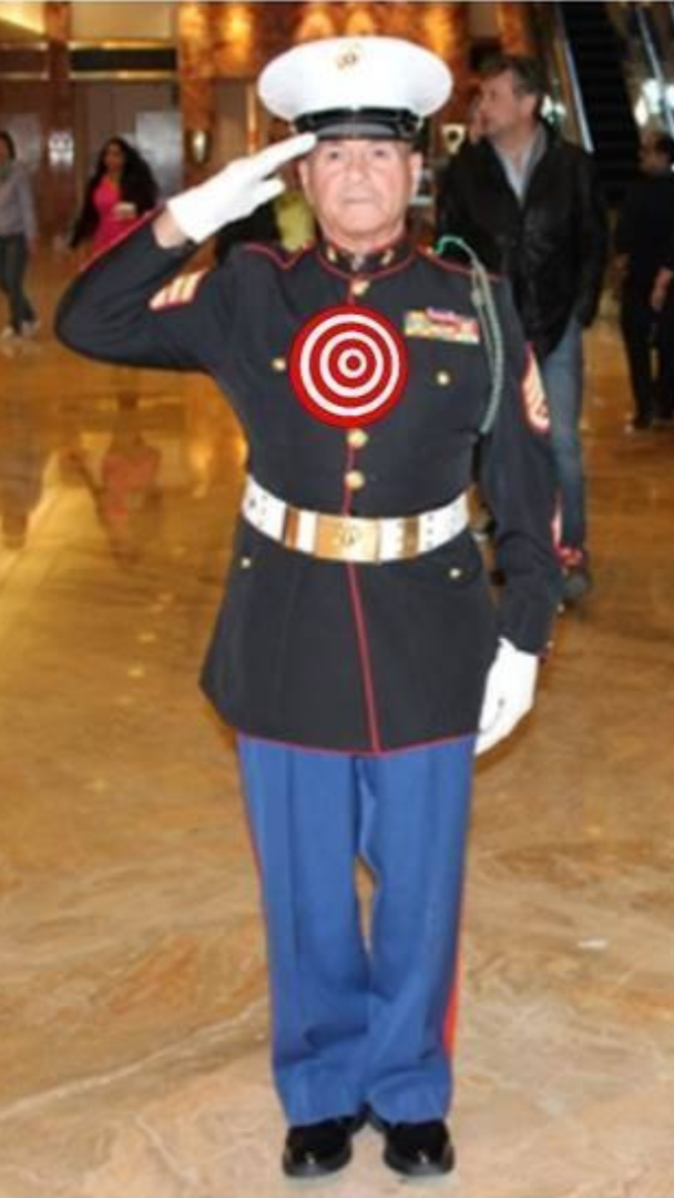 Marine in dress blues, saluting. Red and gold accents, white belt, and blue pants. Indoor setting.