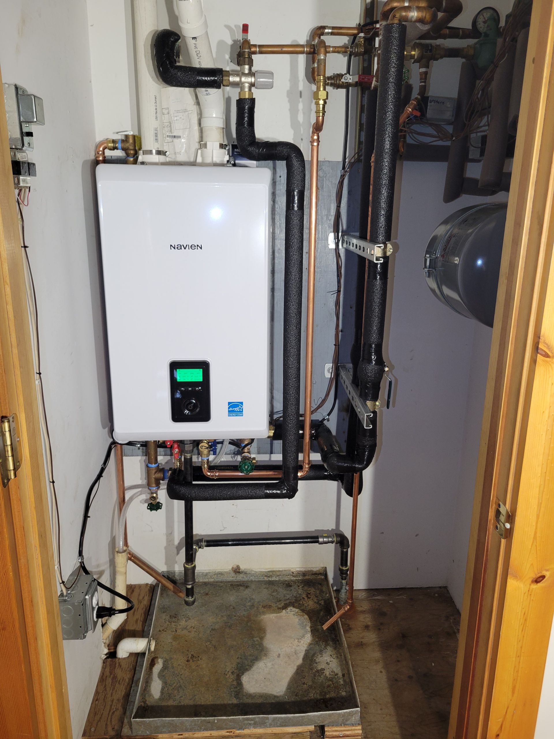 White wall-mounted boiler with black pipes and copper fittings in a small utility room.