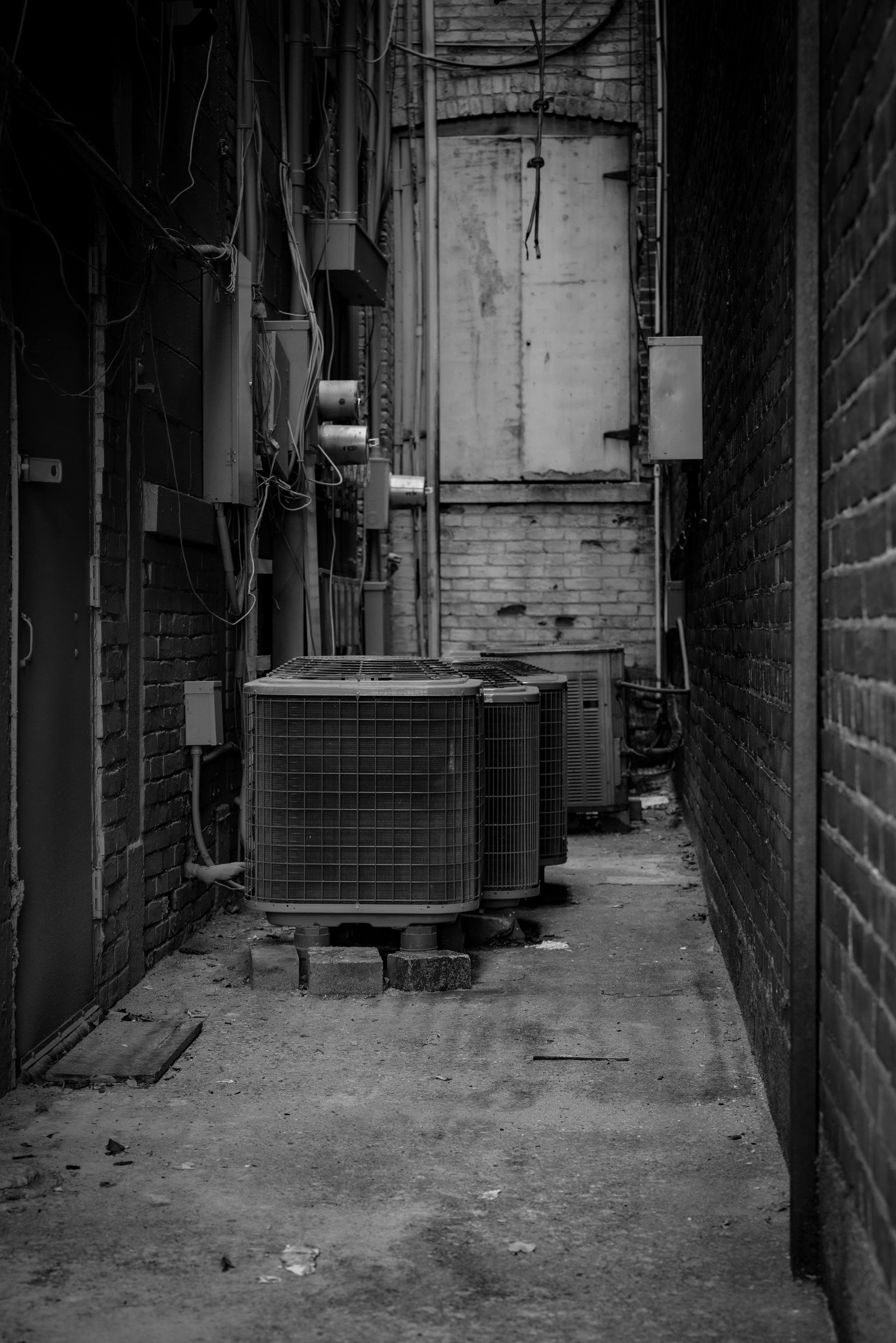 Narrow, brick-walled alleyway with air conditioning units and a metal door. Black and white.