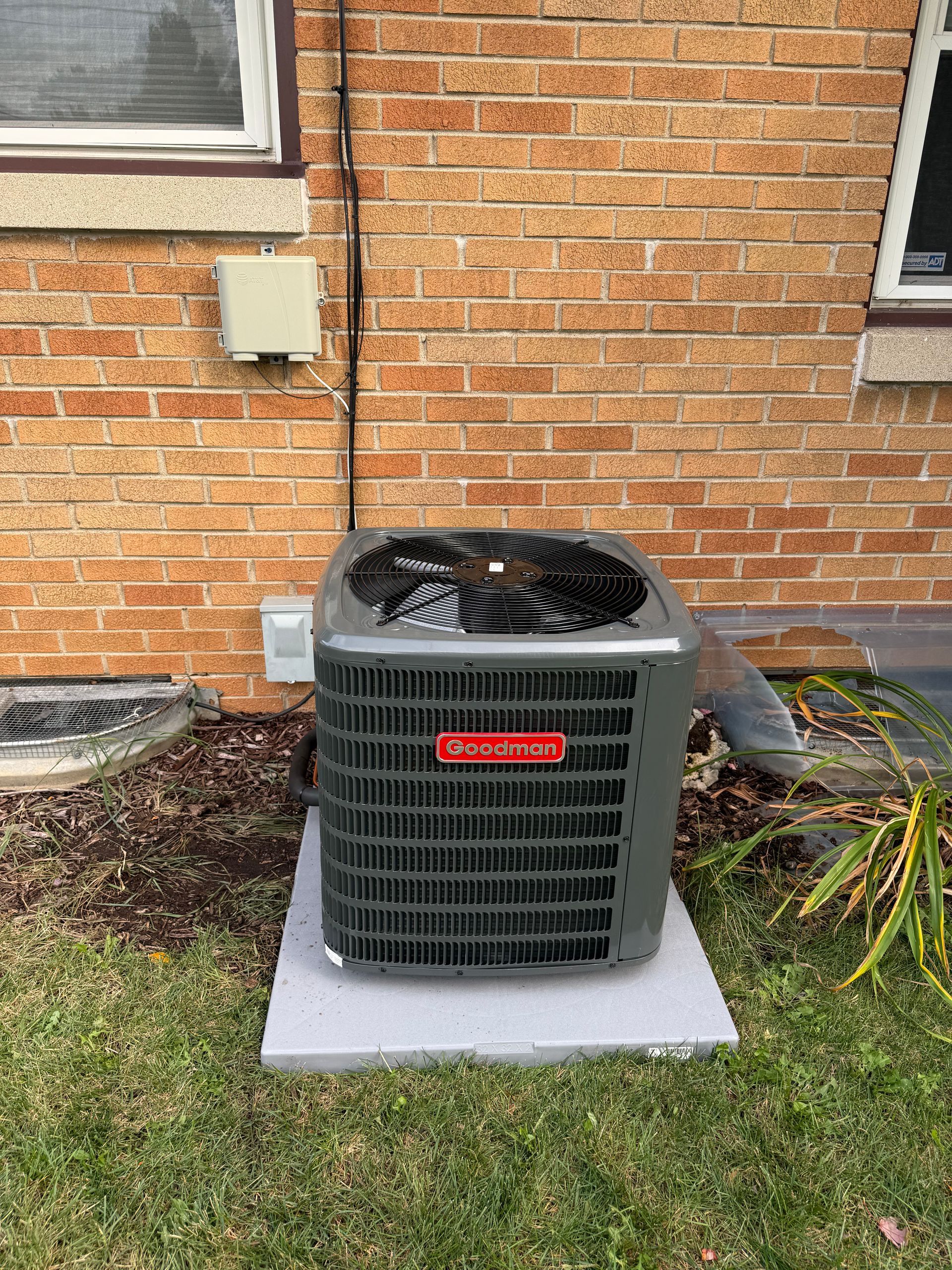 A gray residential air conditioning unit sits on a concrete pad against a brick house wall near a window.