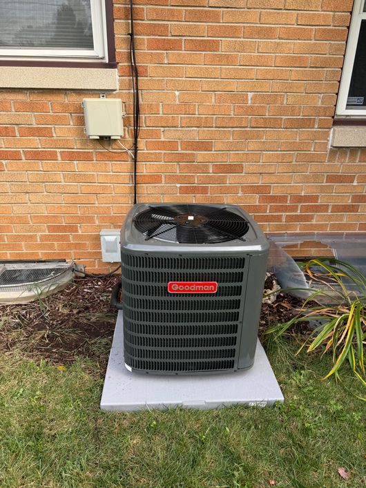 A gray residential air conditioning unit sits on a concrete pad against a brick house wall near a window.