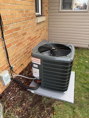 A grey central air conditioning unit sits on a concrete pad next to a brick house exterior.