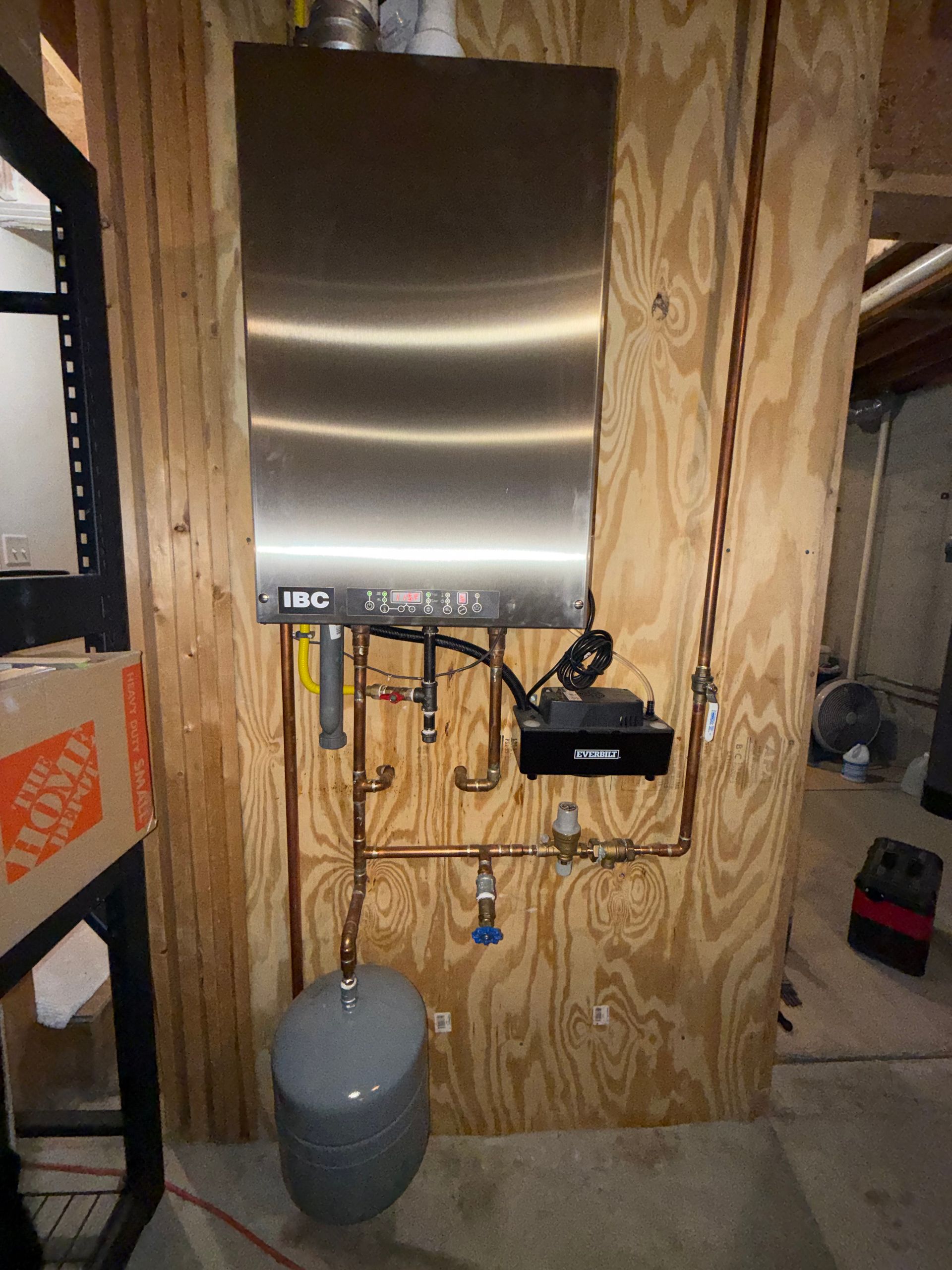 A wall-mounted stainless steel tankless water heater with copper piping, a control unit, and a gray expansion tank.