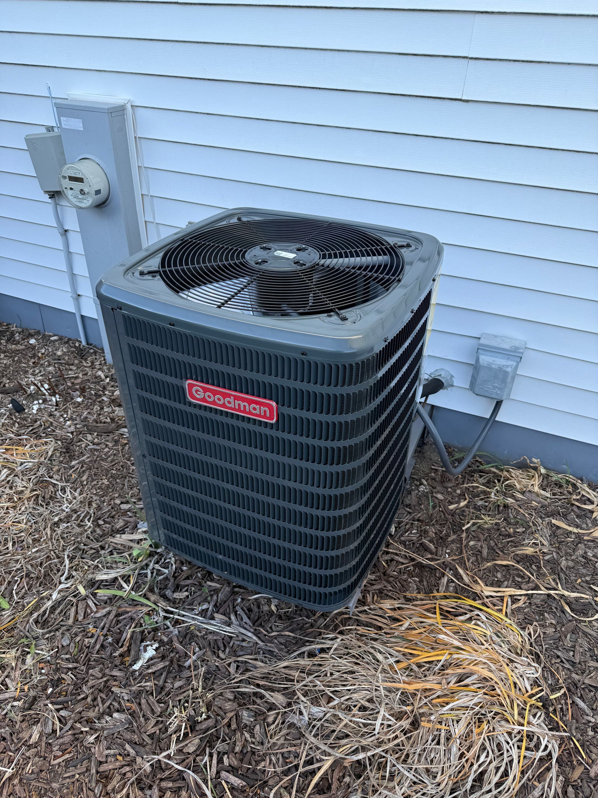 A gray residential air conditioning unit sitting outside against a white siding wall surrounded by brown mulch.